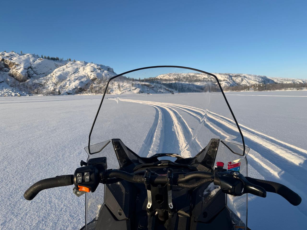View over snowmobile handlebars across a frozen lake with snowy mountains under a blue sky on a snowmobile safari in Kirkenes, Norway