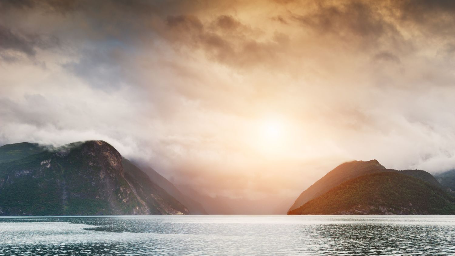 Dramatische Wolken und tiefhängender Nebel tauchen den Himmel über dem Storfjorden in goldenes Licht und erhellen das ruhige Wasser und die majestätischen Berge Westnorwegens.