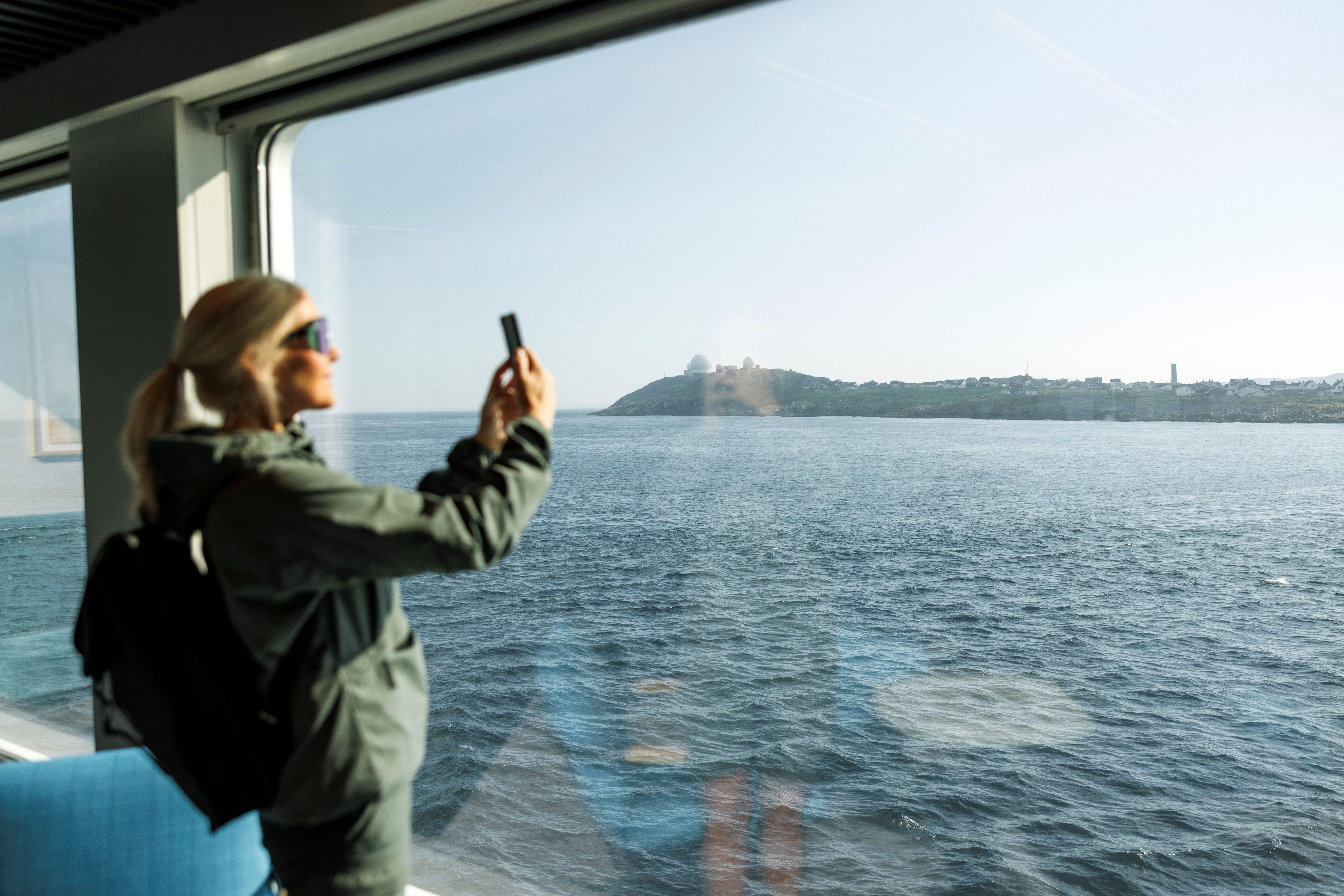 Gjest som fotograferer kystlandskapet som glir forbi, med mobilen gjennom et panoramavindu
