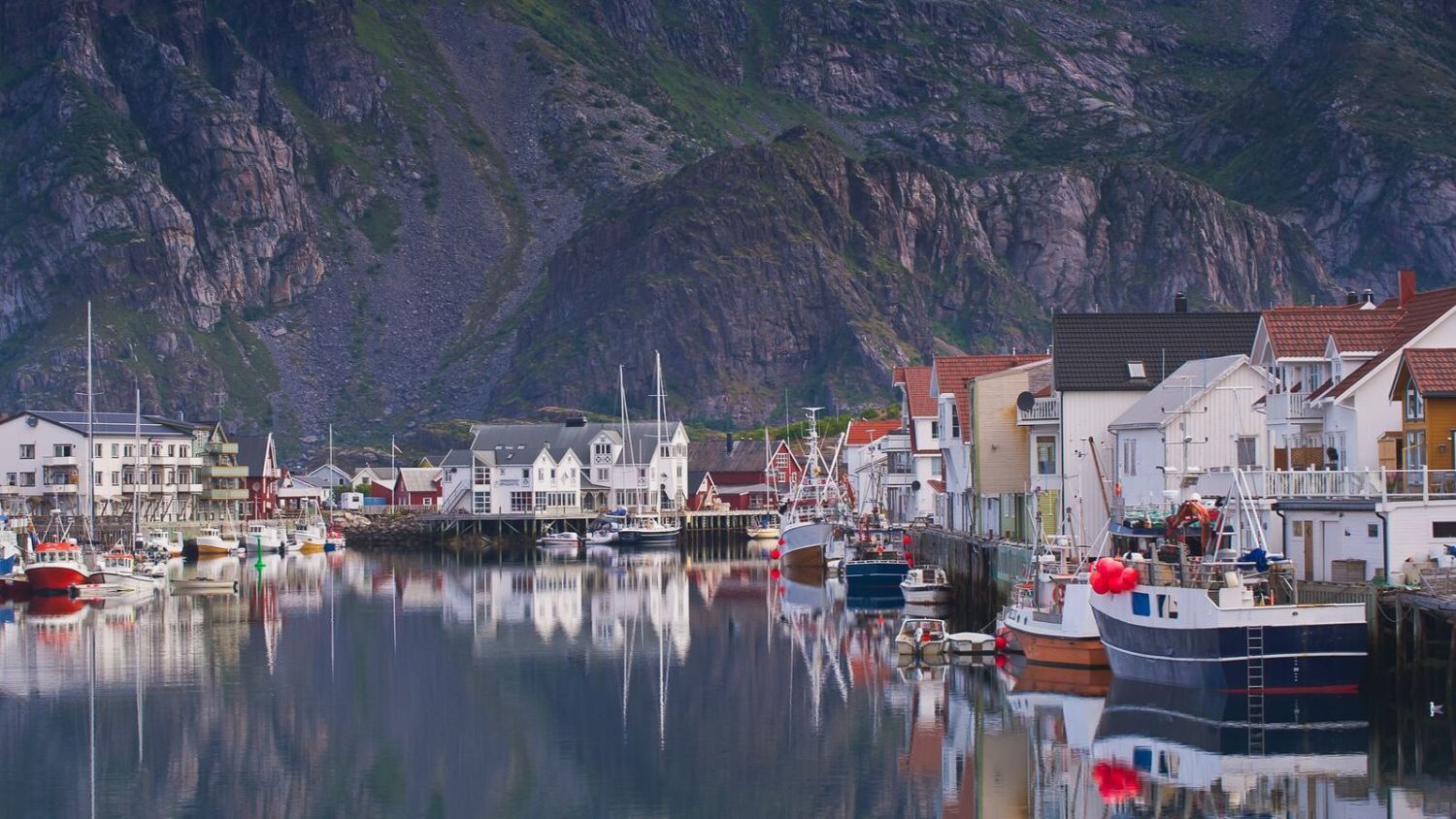Traditioneller Fischerhafen auf den Lofoten mit farbenfrohen Ufergebäuden, vertäuten Fischerbooten und steilen Bergen, die hinter der ruhigen Marina aufragen