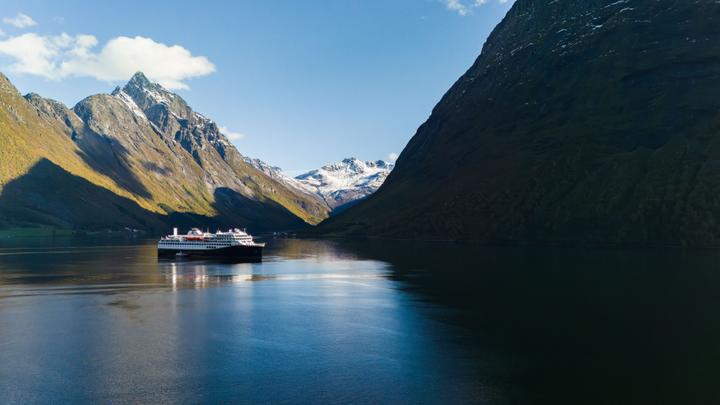 Die Havila Castor fährt durch den Geirangerfjord, umgeben von majestätischen Bergen, und präsentiert die preisgekrönten Küstenreisen von Havila Voyages auf Norwegens ikonischen Fjorden.