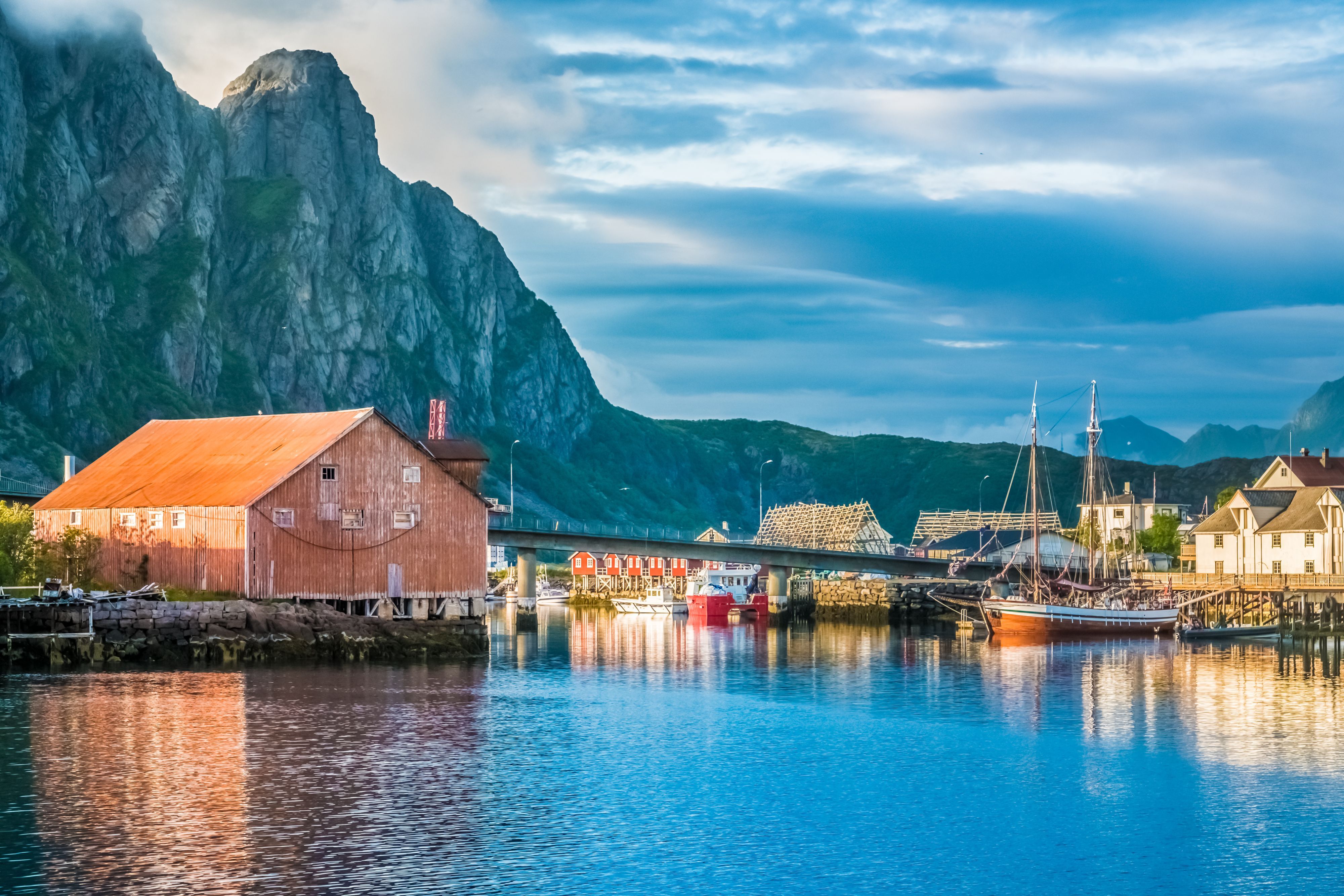 Traditionelles Fischerdorf und Hafen in Reine auf den Lofoten in Norwegen, mit farbenfrohen Gebäuden an der Uferpromenade, Holzbooten, dramatischen Granitbergen und ruhigem, spiegelglattem Wasser