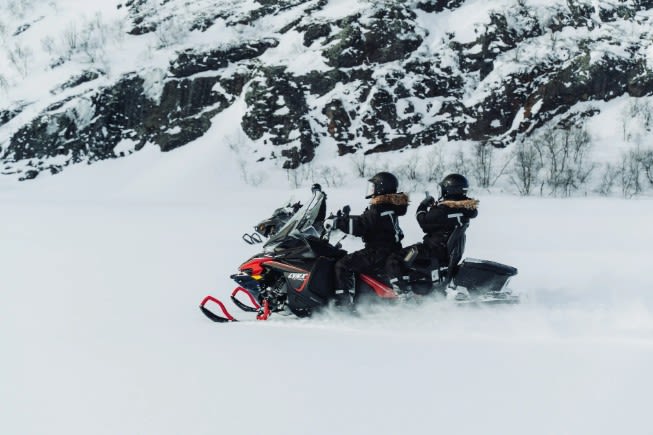 Two helmeted riders on a snowmobile speeding across snow with rocky mountains behind on a snowmobile safari in Kirkenes, Norway