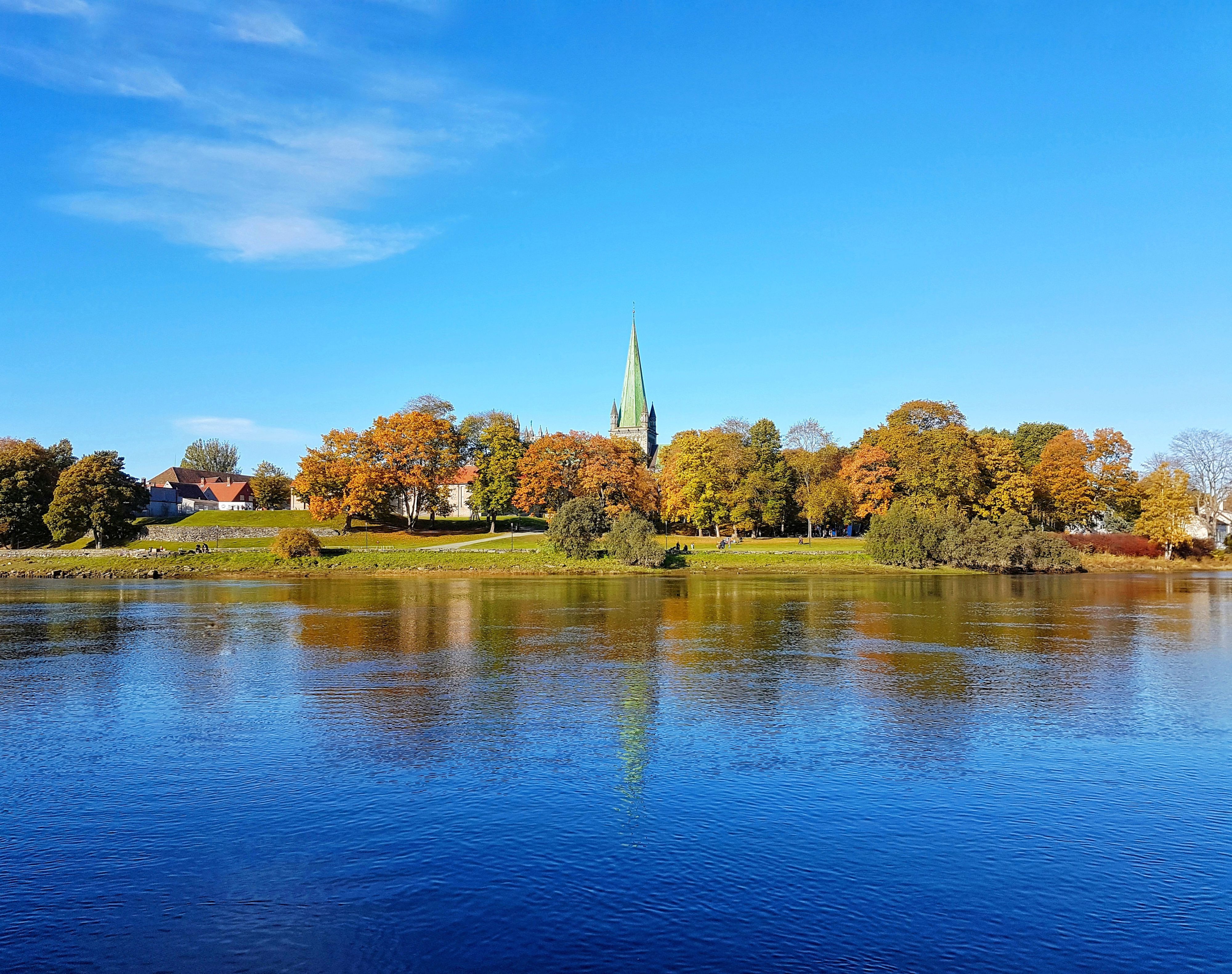 Der Turm des Nidarosdoms und herbstliche Bäume spiegeln sich im Fluss Nidelva in Trondheim, Norwegen