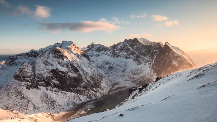 Schneebedeckte Berggipfel und ein zugefrorener Fjord, die im goldenen Februarlicht entlang Norwegens dramatischer arktischer Küste leuchten