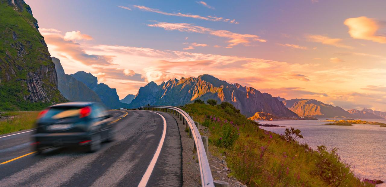 En bil kjører langs en naturskjønn kystvei på Lofoten ved solnedgang, med dramatiske fjell, rolig hav og livlige sommerfarger som skaper en uforglemmelig norsk biltur.