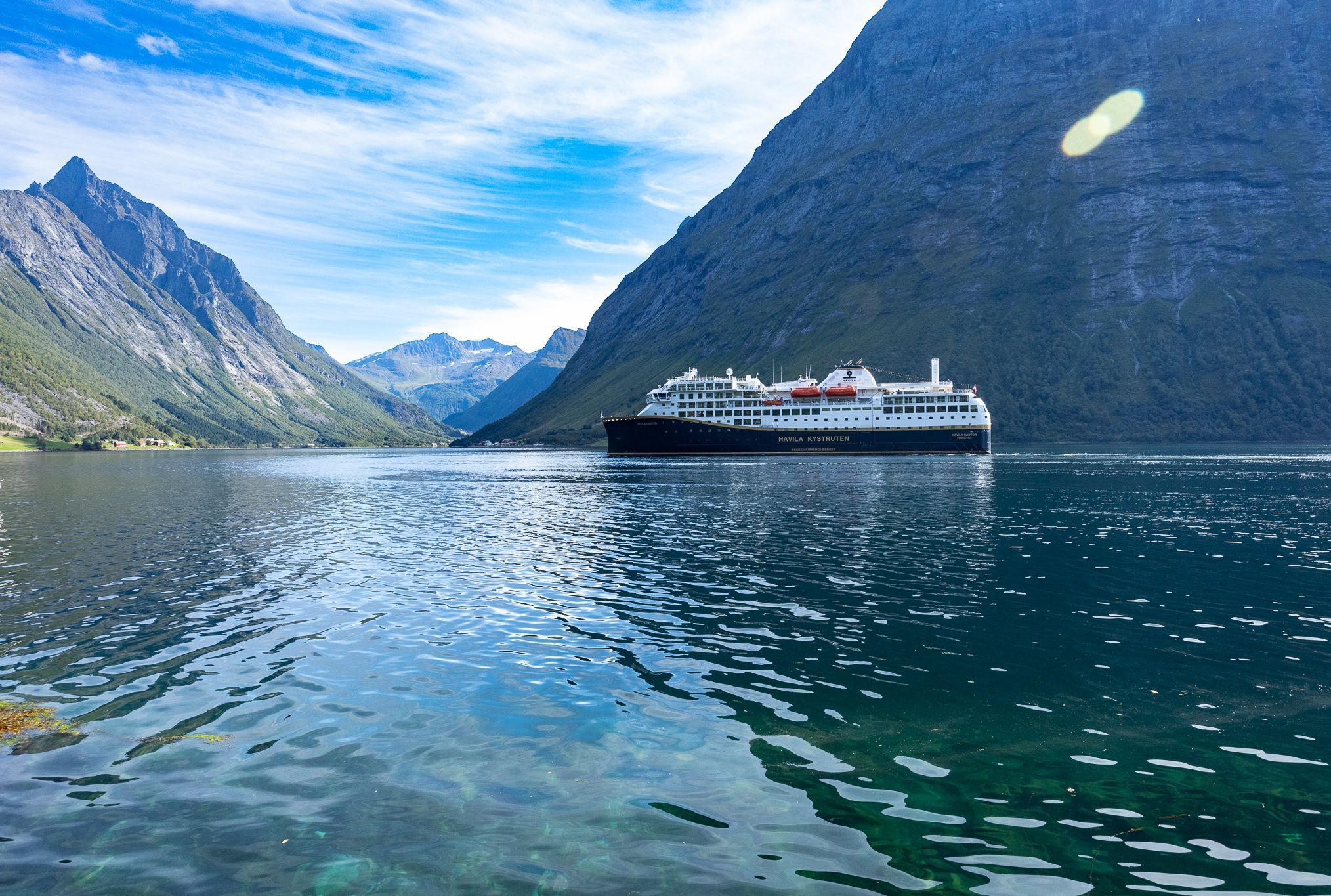Havila kryssningsfartyg som seglar genom Hjørundfjorden, omgivet av höga berg och kristallklart vatten på sommaren.