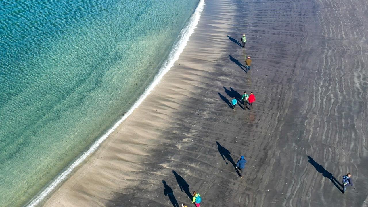 Flyfoto av mennesker som går langs en arktisk kyst på en sandstrand med klart turkis vann i Norge