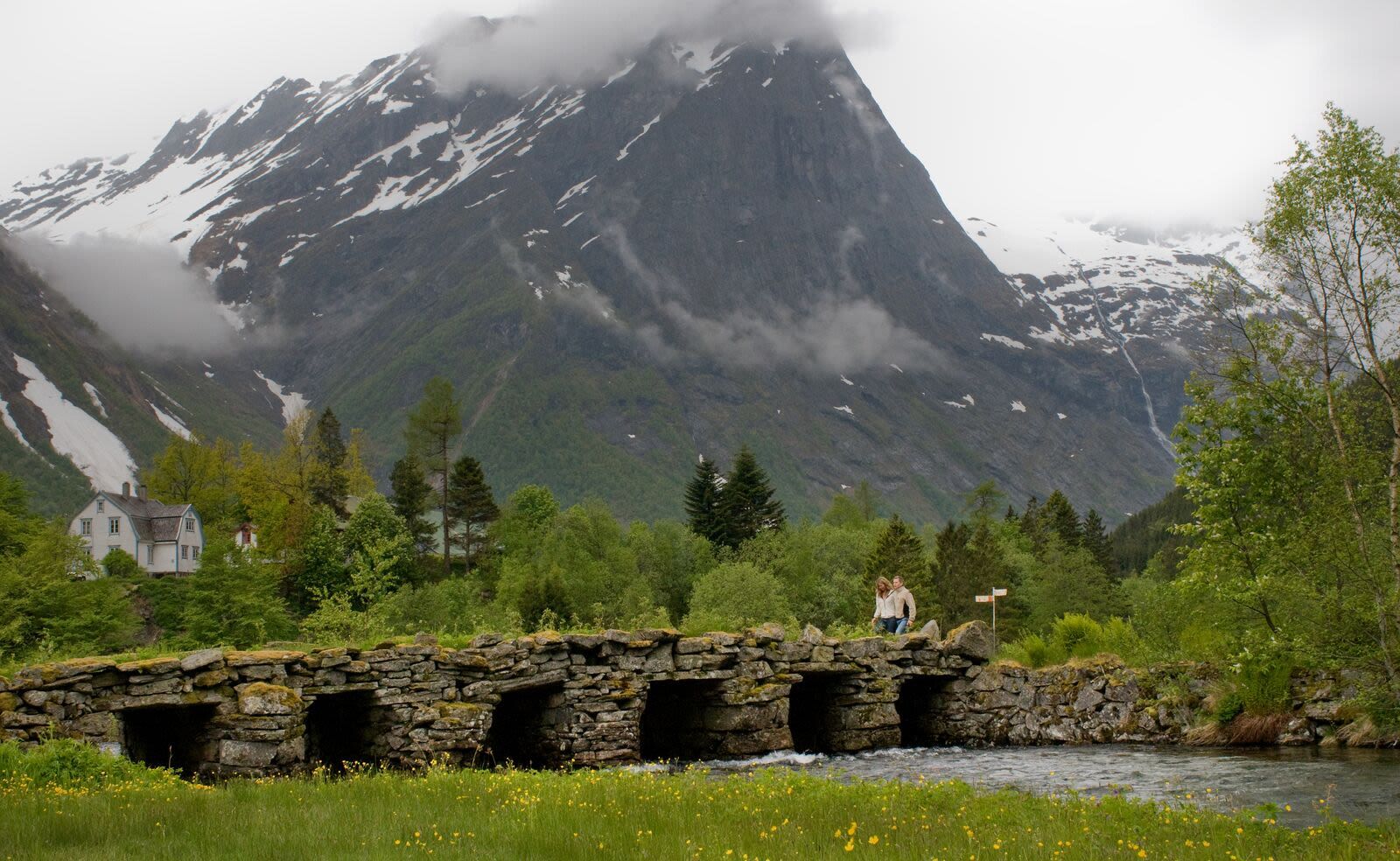 Couple walking across an old stone arch bridge over a river below cloudy mountains in rural Norway