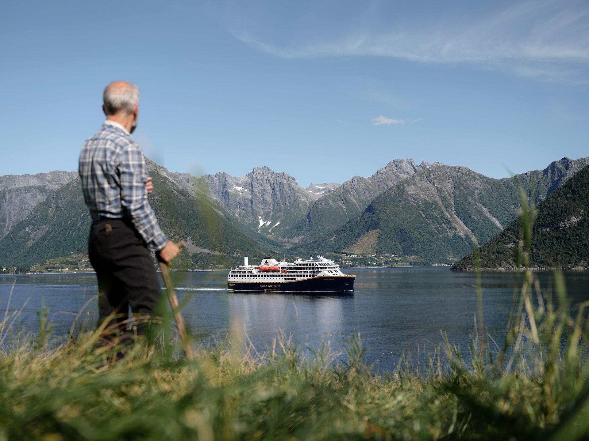 Ein Einheimischer beobachtet, wie ein Schiff von Havila Voyages durch einen friedlichen norwegischen Fjord fährt, umgeben von majestätischen Bergen und einer atemberaubenden Küstenlandschaft.