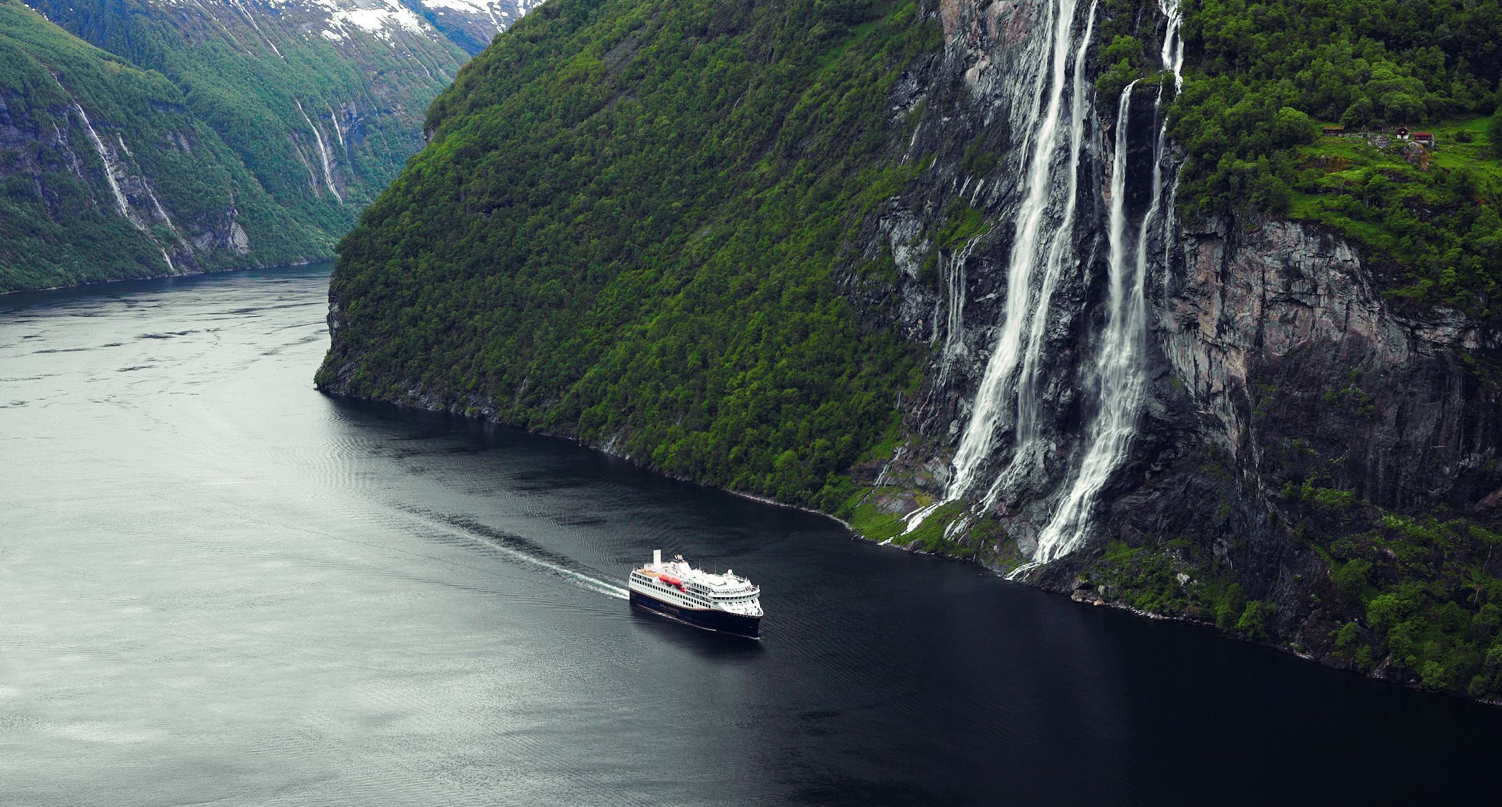 Havila Voyages kystcruiseskip seiler gjennom Geirangerfjorden under mektige fossefall og frodige fjellklipper i Norge.