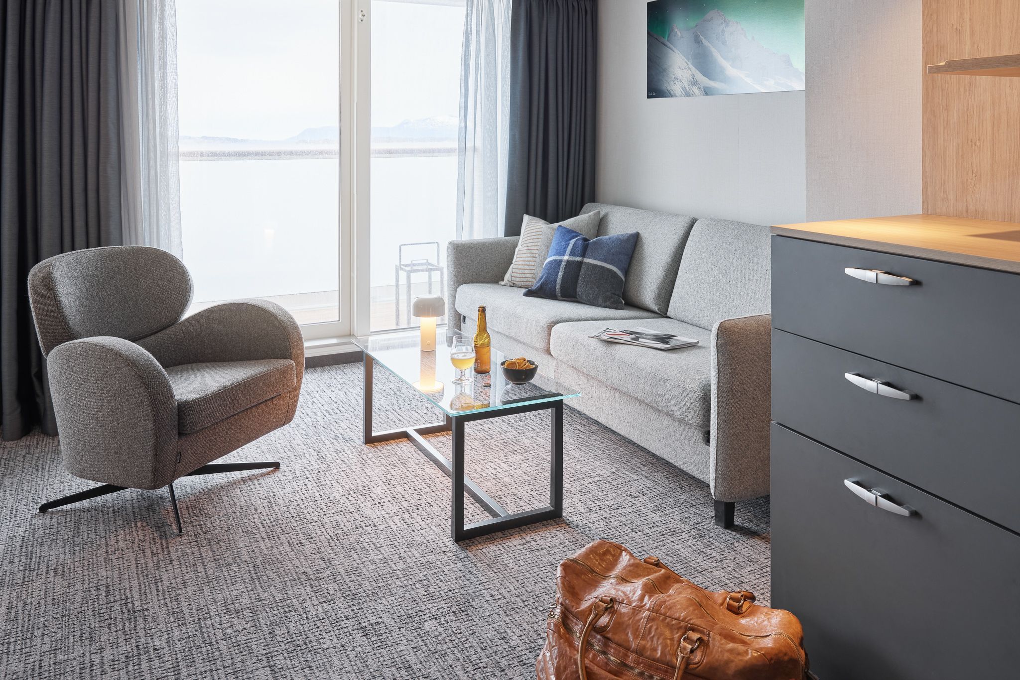 Tilgjengelig juniorsuite med stuedel som har en komfortabel sofa, lenestol, glassbord og gulv-til-tak-dører som åpner ut mot en privat balkong med havutsikt.