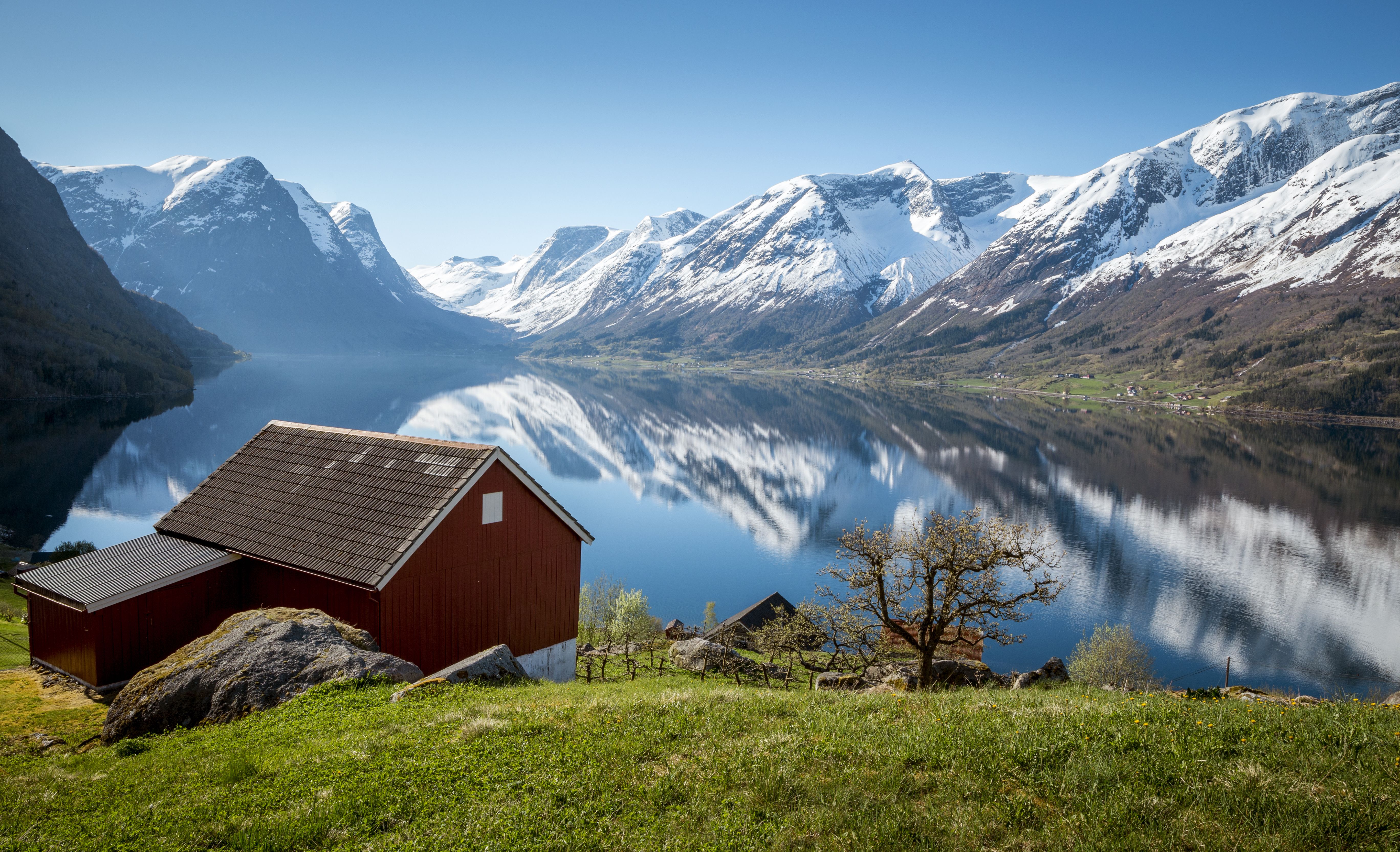 Rødt båthus i tre på grønt gress ved en stille fjord, med snødekte fjell som speiler seg i vannet