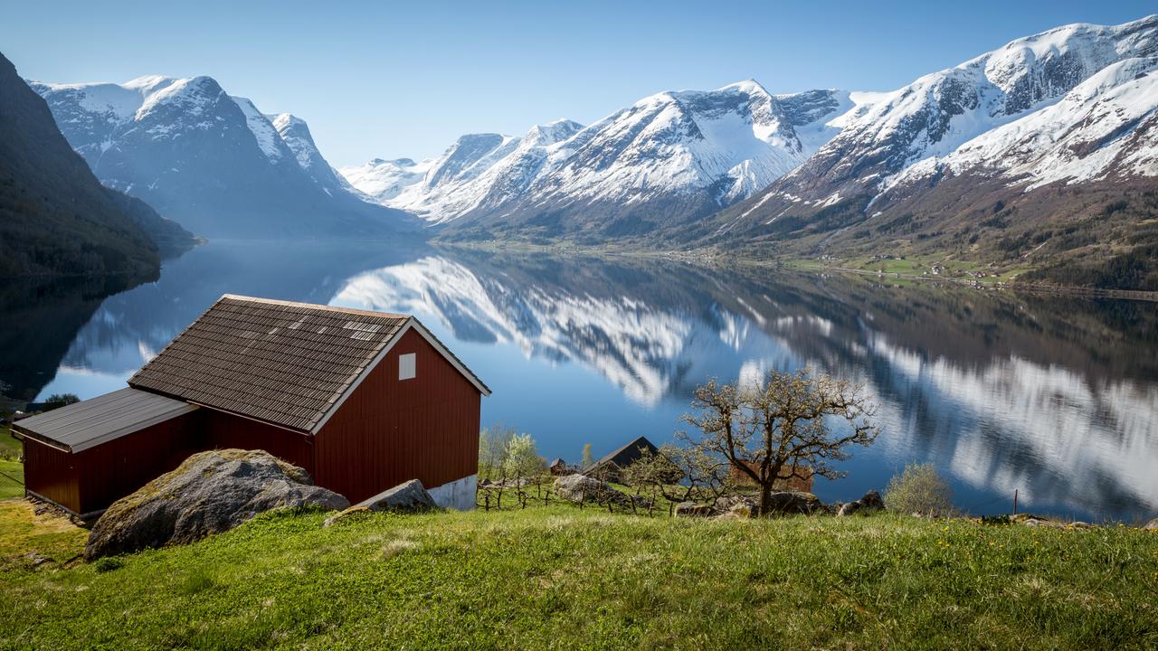 Rött båthus i trä på grönt gräs ovanför en spegelblank fjord, med snöklädda berg som speglas i vattnet