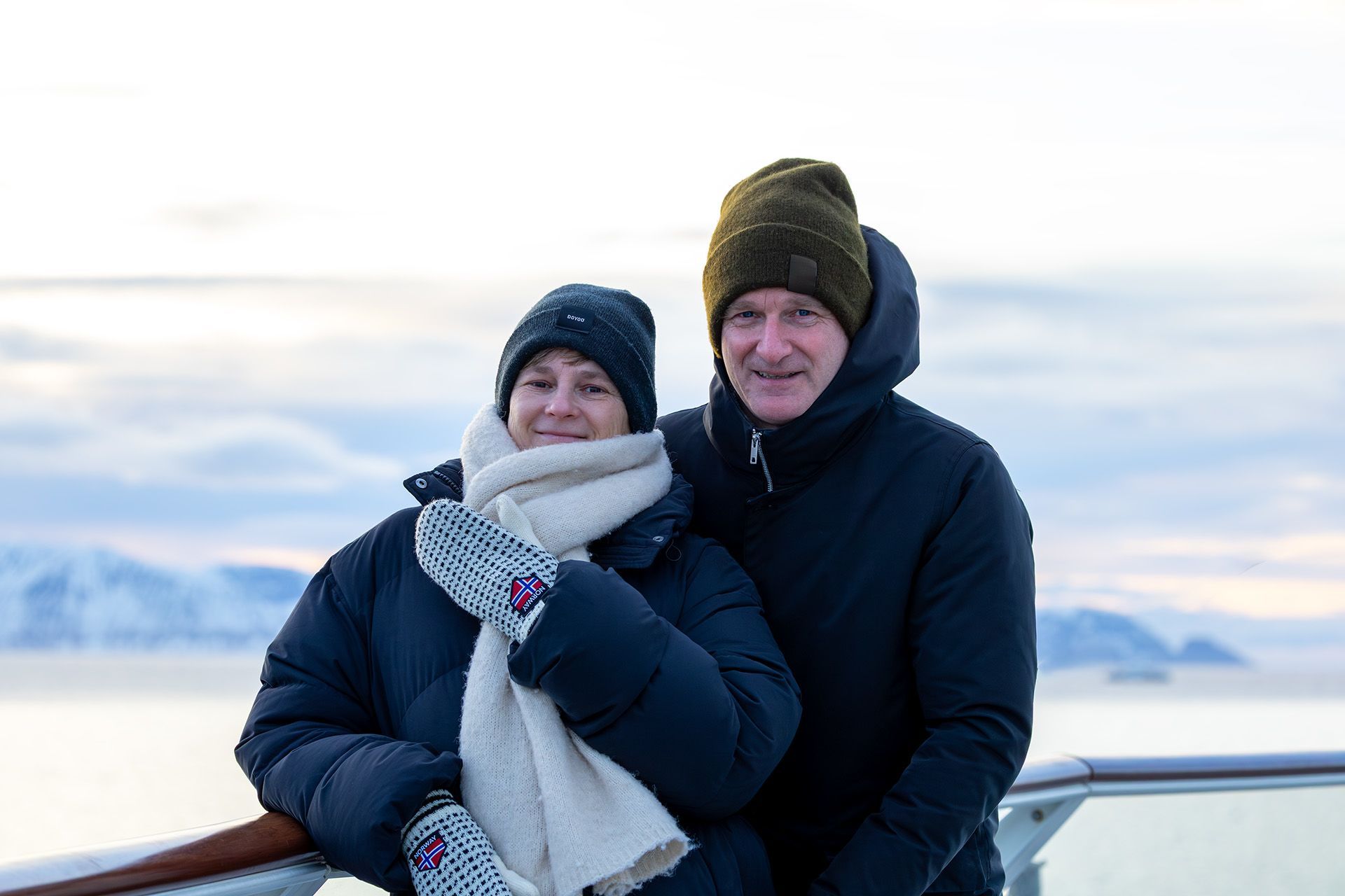 Ein Paar, warm gekleidet, steht auf dem Außendeck eines Havila-Kystruten-Schiffs und genießt die arktische Winterlandschaft mit schneebedeckten Bergen und ruhigen Küstengewässern im Hintergrund.
