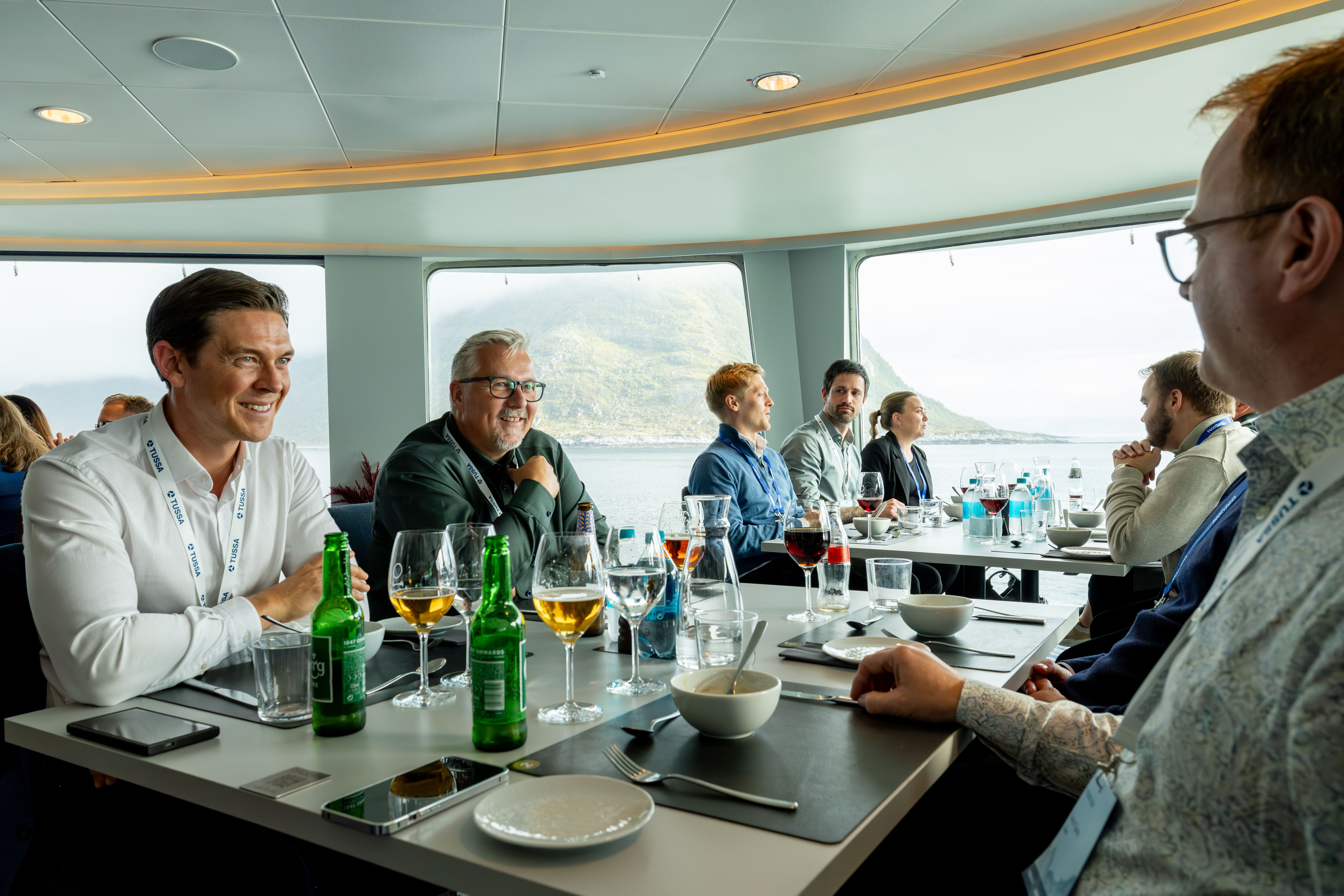 Gäster samtalar runt ett dukat matbord intill panoramafönster ombord