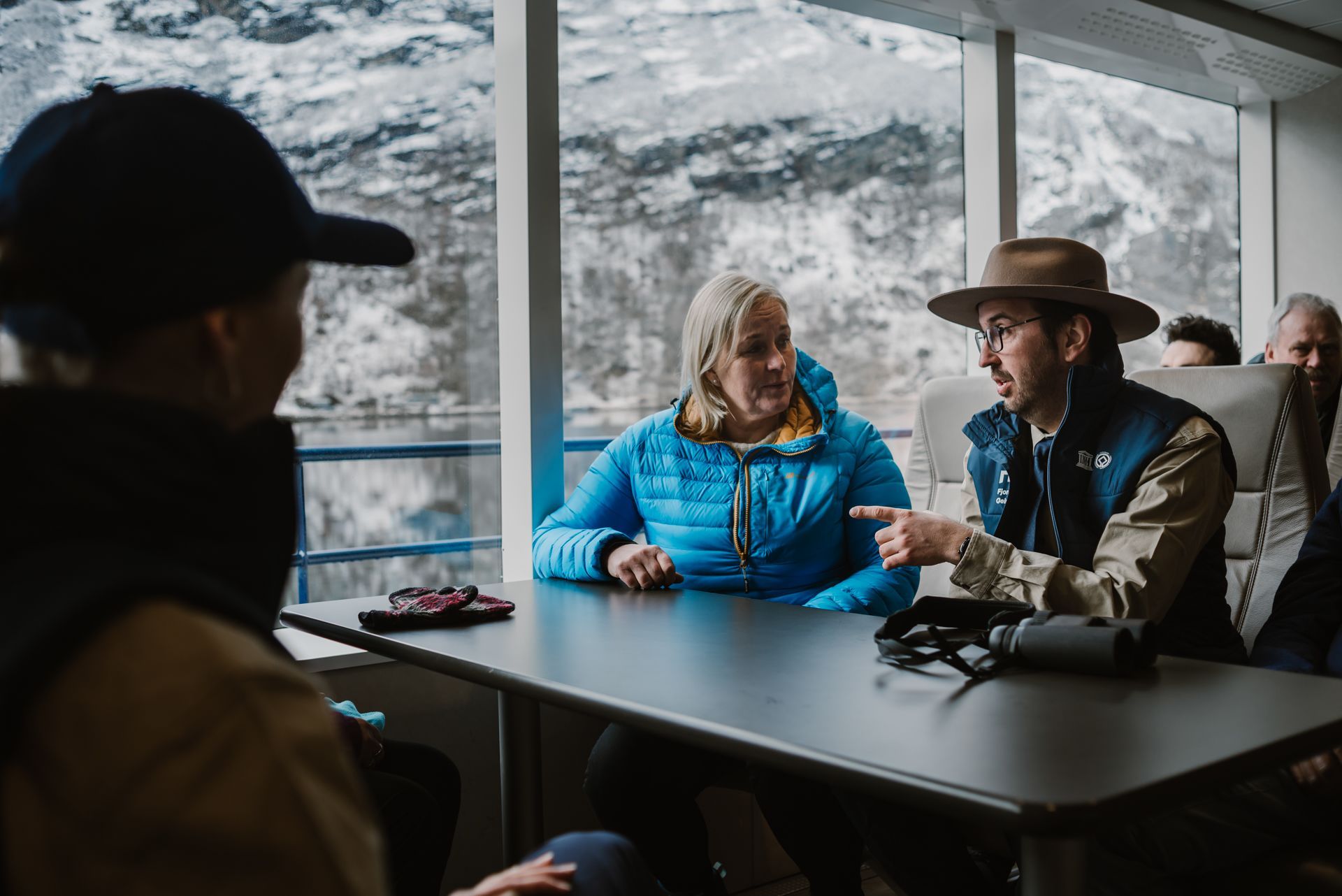 Fjord cruise guide talking with passengers inside the boat cabin with snowy mountains outside, Geiranger, Norway