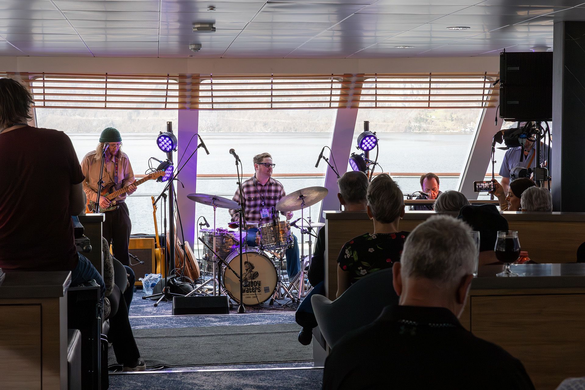 Bill Troiani opptrer live med bandet sitt i panoramasalongen om bord på Havila Voyages Blues Cruise 2027, mens gjestene nyter konserten og den vakre utsikten over fjorden gjennom de store vinduene.