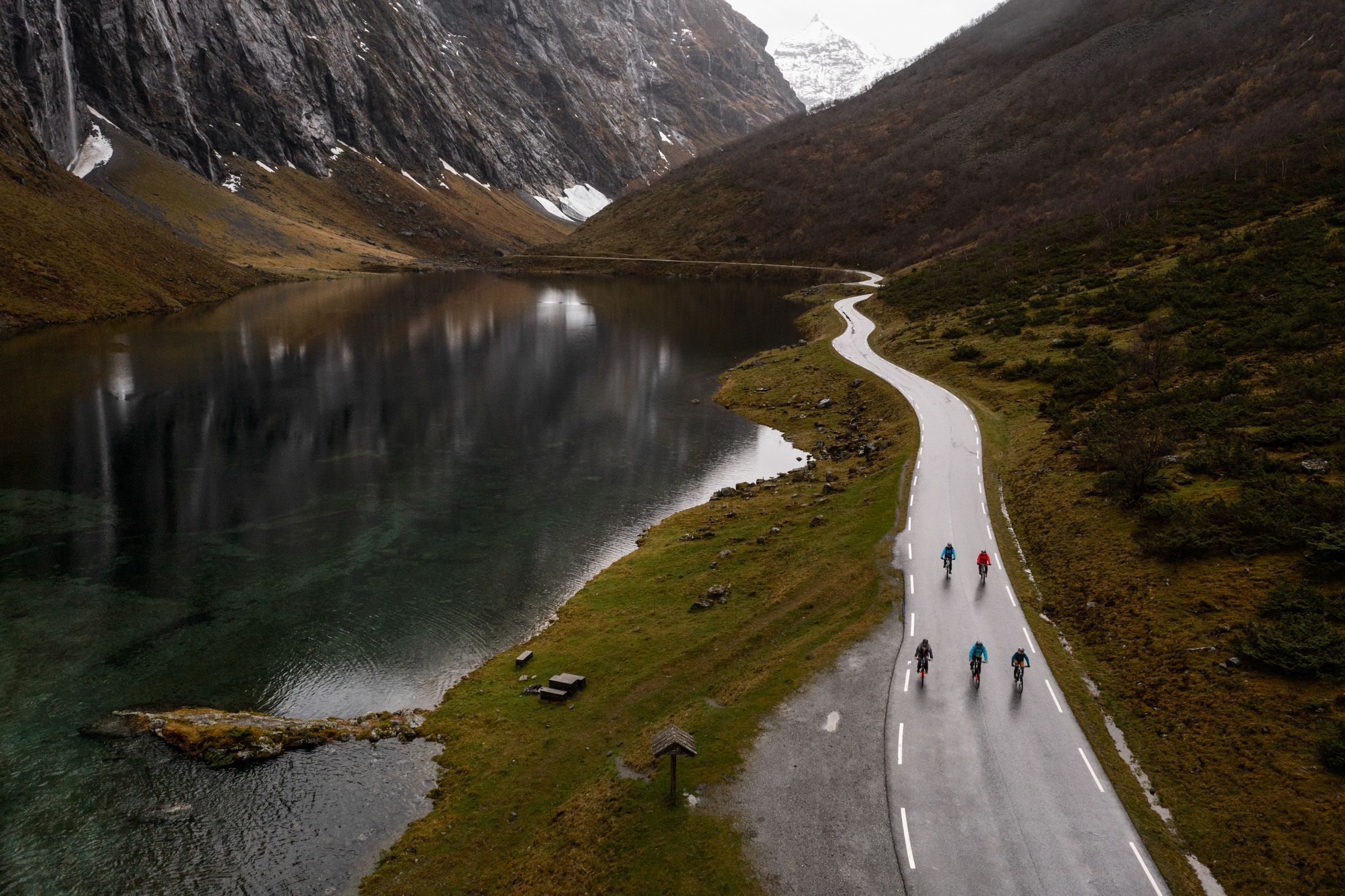 Aerial view of cyclists riding a winding road beside a calm mountain lake near Hjørundfjorden, Norway