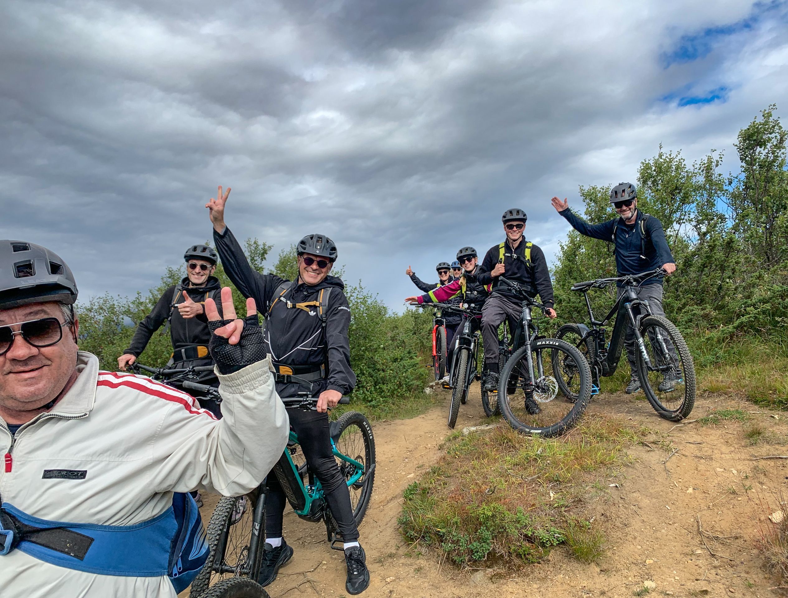 Glad gruppe syklister som tar en selfie og vinker med elsyklene sine på en fjellsti under en elsykkeltur i Bodø, Norge