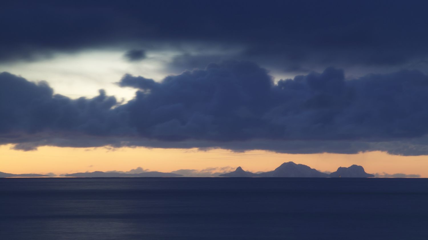 Dunkle Sturmwolken über dem Vestfjord bei Sonnenuntergang, mit fernen Bergsilhouetten, die vor dem farbenprächtigen Abendhimmel in Nordnorwegen leuchten.