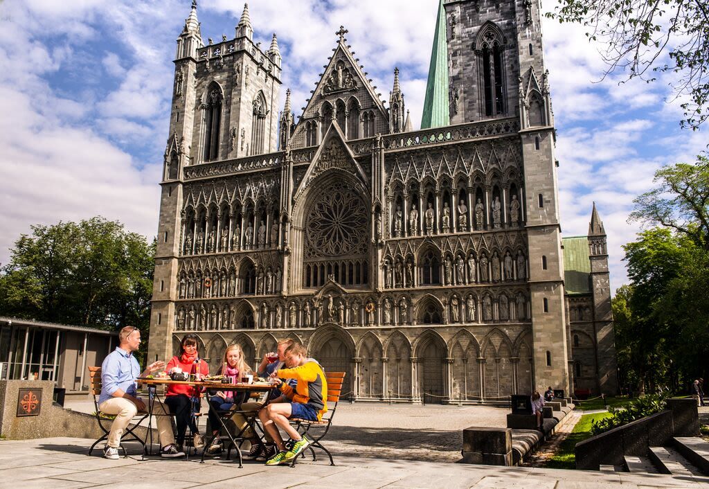 Besucher genießen die Außenterrassen vor dem Nidarosdom in Trondheim, Norwegen, mit Blick auf die ikonische gotische Fassade, die kunstvollen Steinmetzarbeiten und die historische mittelalterliche Architektur.