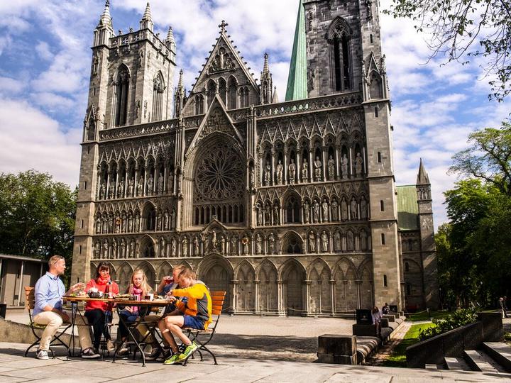 Besucher genießen die Außenterrassen vor dem Nidarosdom in Trondheim, Norwegen, mit Blick auf die ikonische gotische Fassade, die kunstvollen Steinmetzarbeiten und die historische mittelalterliche Architektur.