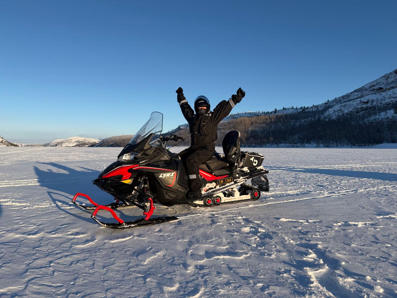 Excited rider with arms raised on a snowmobile on a frozen lake with snowy mountains under a blue sky on a snowmobile safari in Kirkenes, Norway