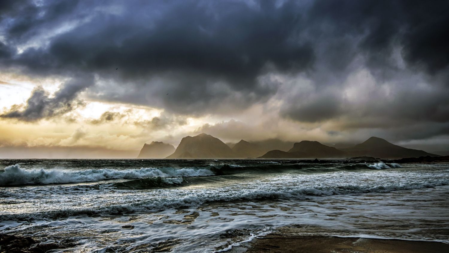 Dunkle Sturmwolken ziehen über den Vestfjord in Nordnorwegen auf, während mächtige Wellen auf die Küste zurollen und sich in der Ferne die Silhouetten zerklüfteter Berge abzeichnen.