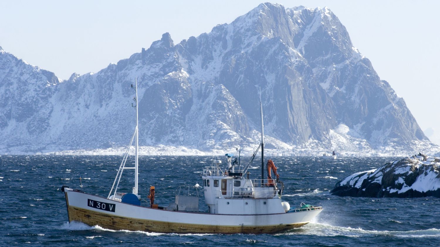 Traditionelles norwegisches Fischerboot, das über blaues Küstenwasser segelt, während sich im Hintergrund die schneebedeckten Lofoten-Berge dramatisch unter einem klaren Winterhimmel erheben