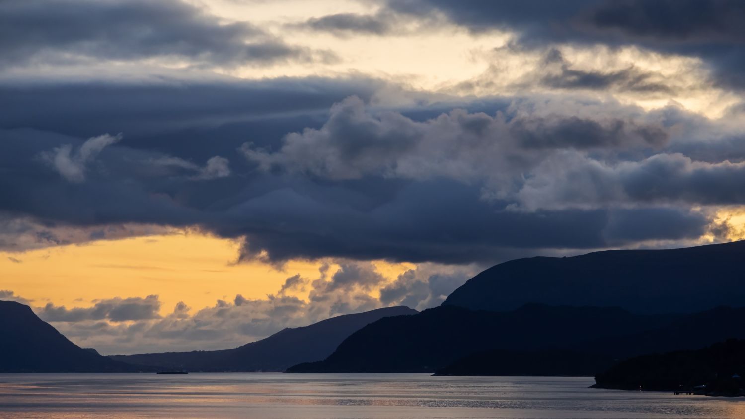 Dramatischer Sonnenuntergang über dem Storfjorden in Westnorwegen, mit dunklen Bergsilhouetten, ruhigem Fjordwasser und dramatischen Sturmwolken, die vom goldenen Abendlicht erleuchtet werden.