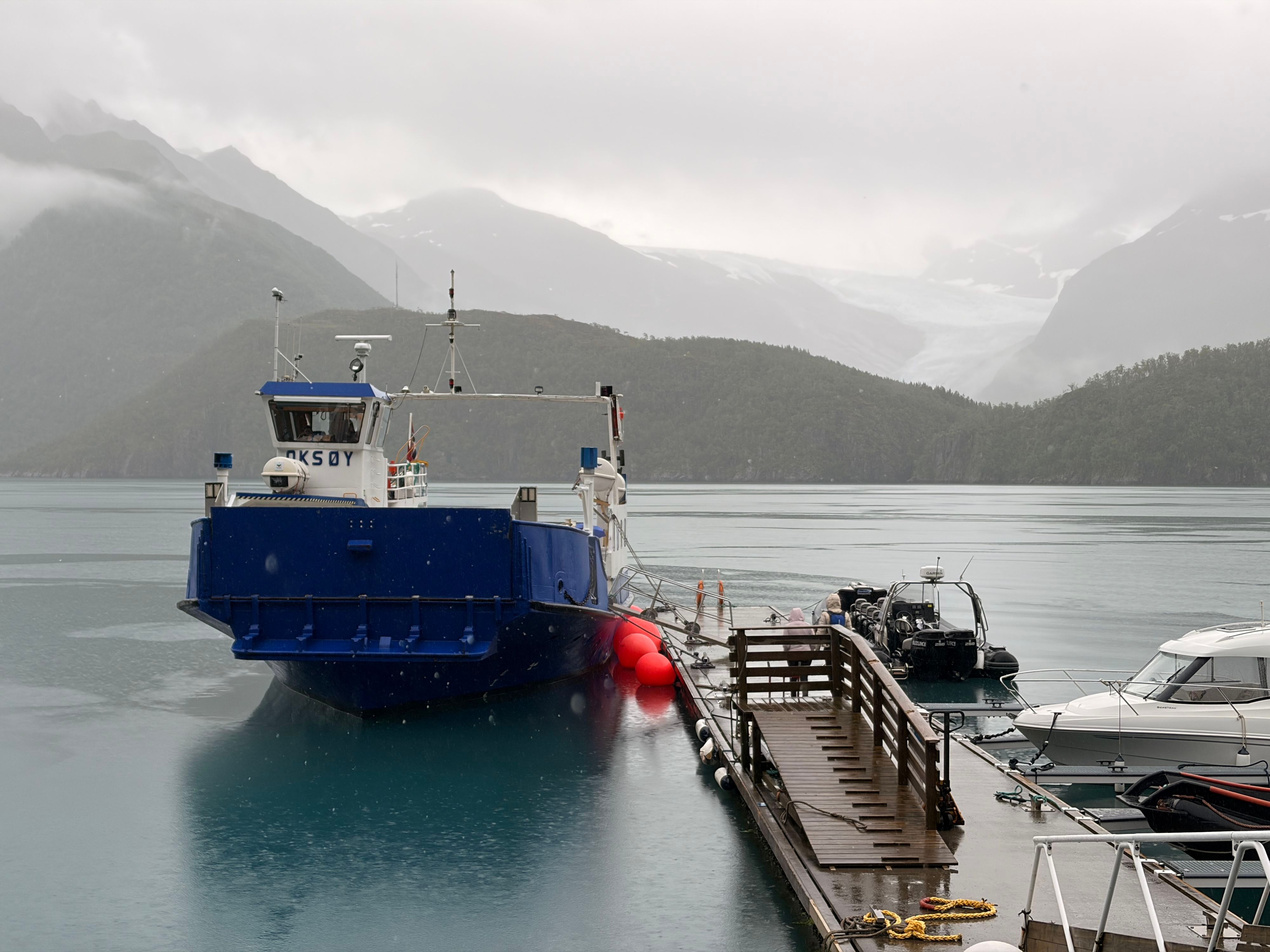 Blau-weiße Fähre, die bei Regen am Kai des Holandsfjords liegt, während im Hintergrund ein Ausläufer des Svartisen-Gletschers am Berghang zu sehen ist