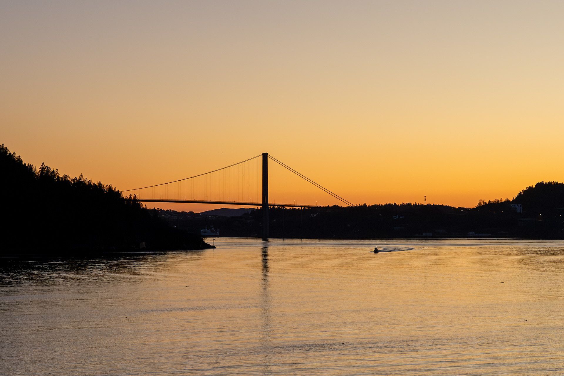 Suspension bridge silhouetted against an orange sunset sky over calm water