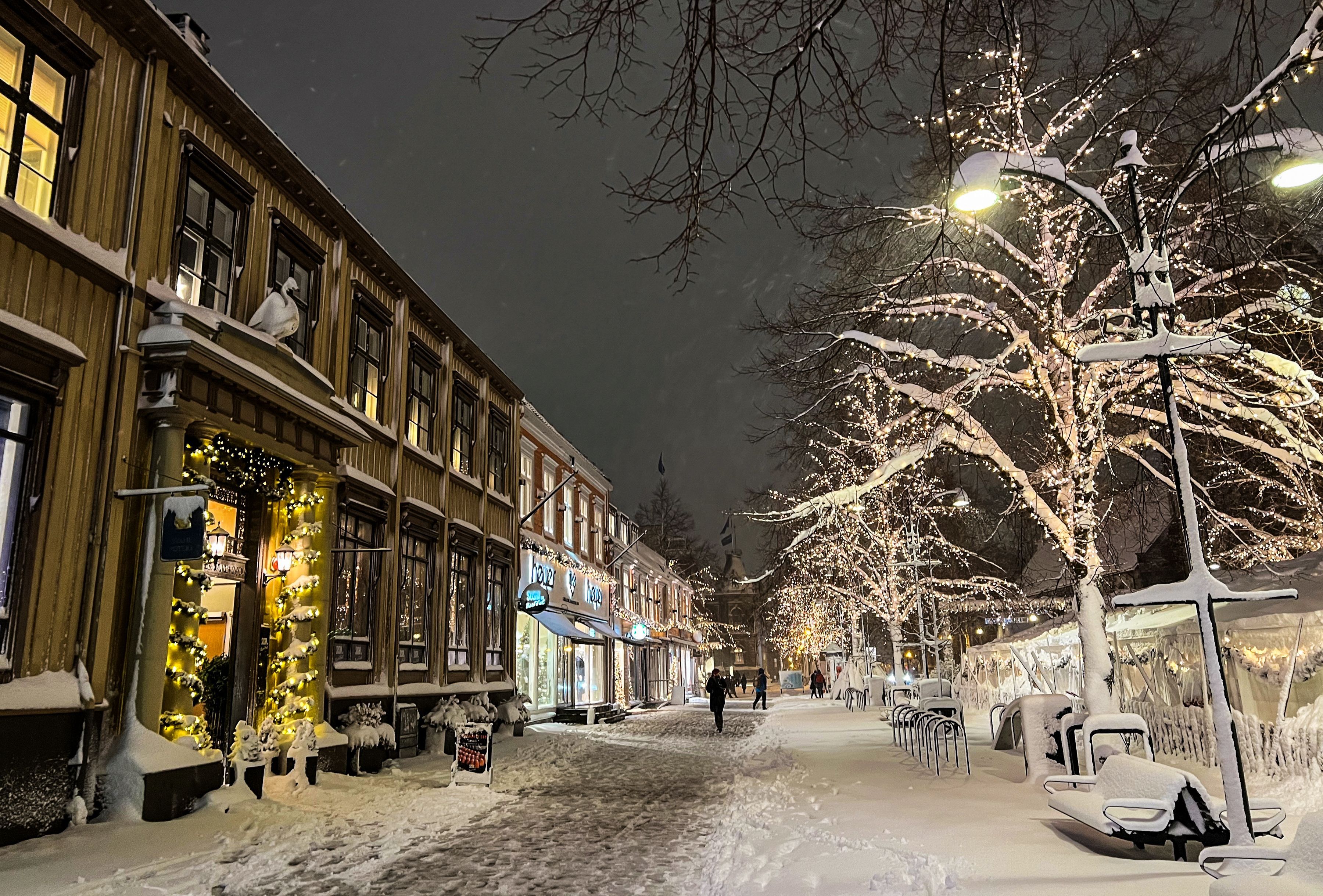 TrondheimSnow-covered street in Trondheim decorated with Christmas lights, historic wooden buildings, and people walking through a festive winter evening.