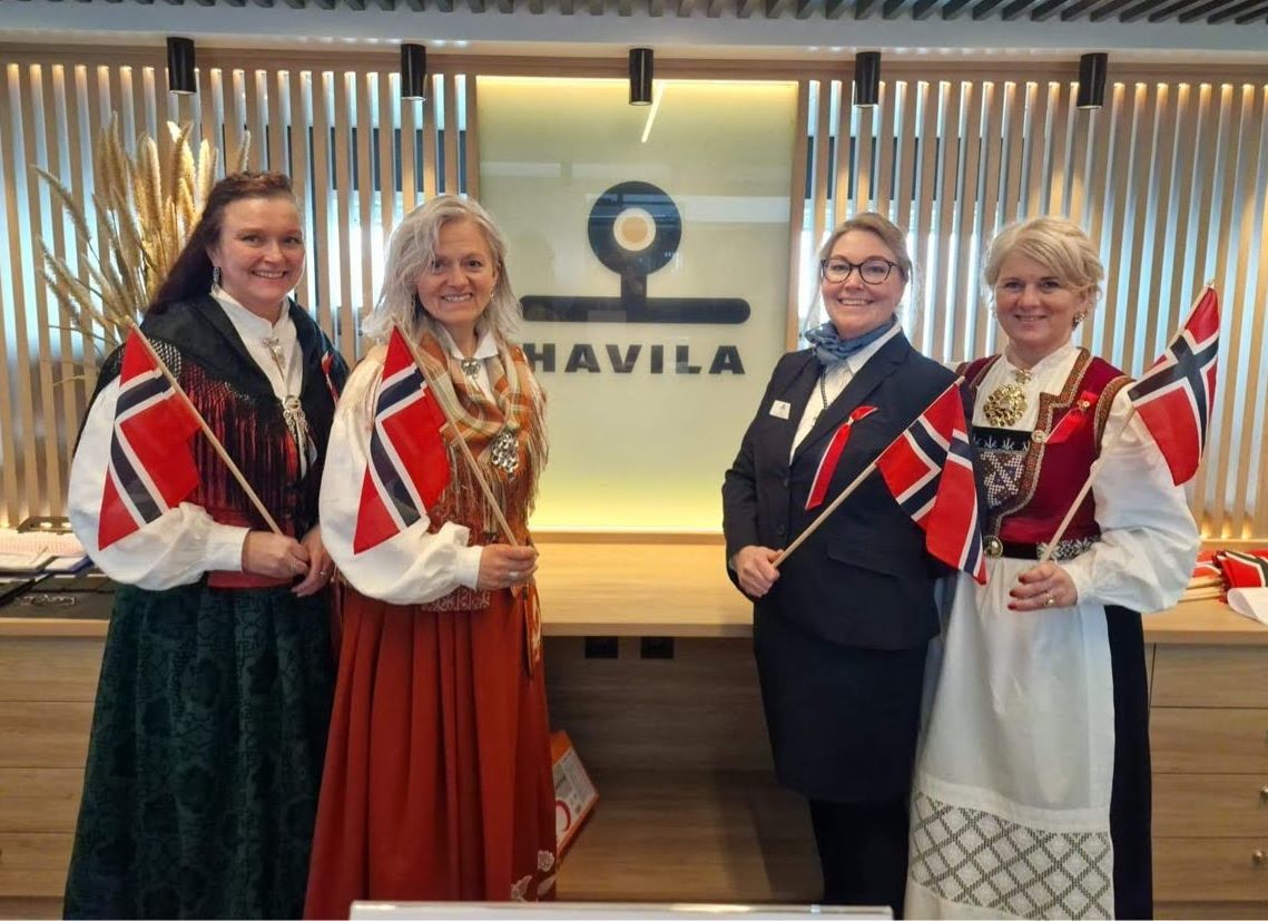 Havila crew members celebrating Norway's Constitution Day (17 May) in traditional bunads, holding Norwegian flags aboard a Havila Kystruten ship