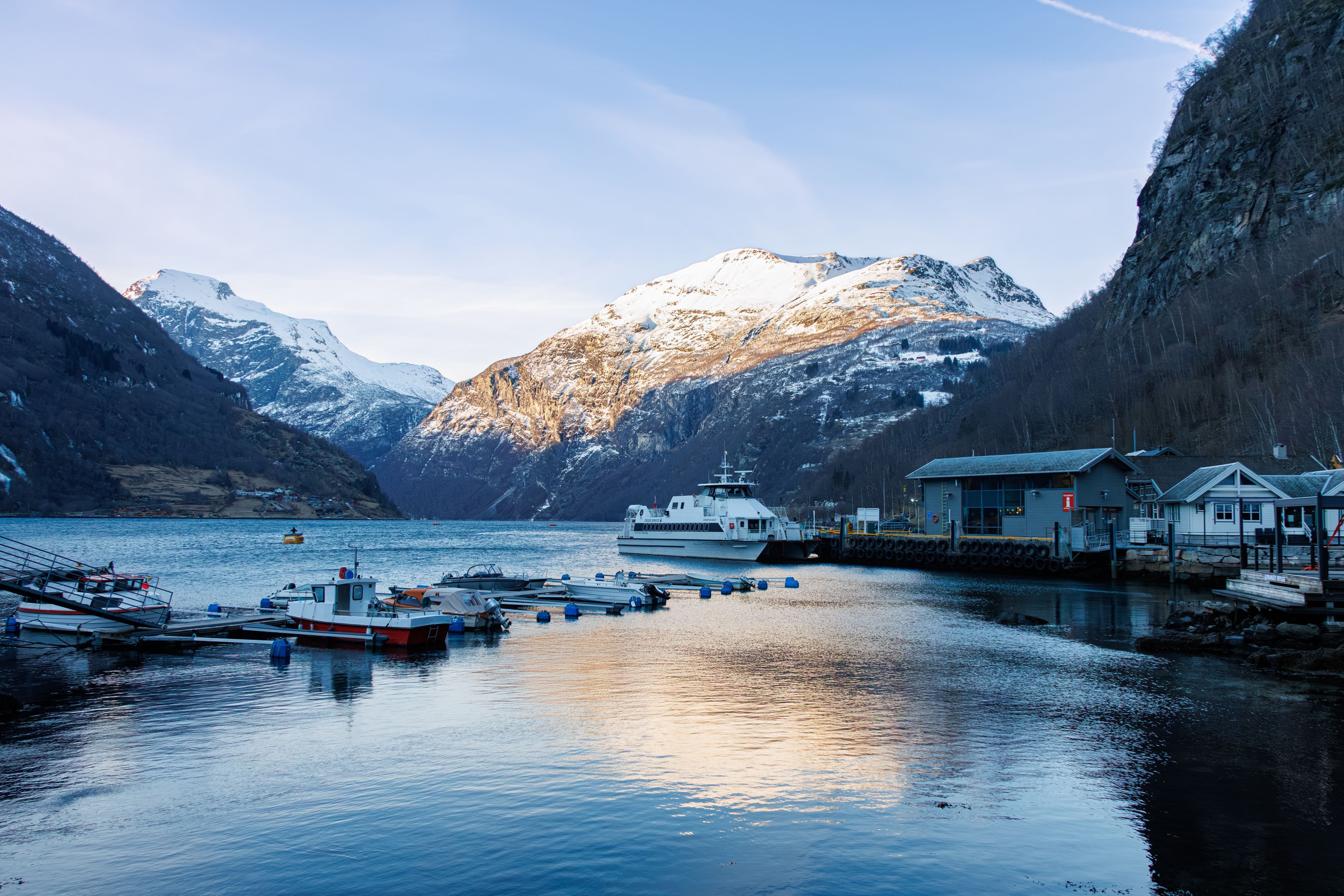 Geiranger harbour with a cruise boat and small boats below snow-capped mountains at winter sunset, Norway
