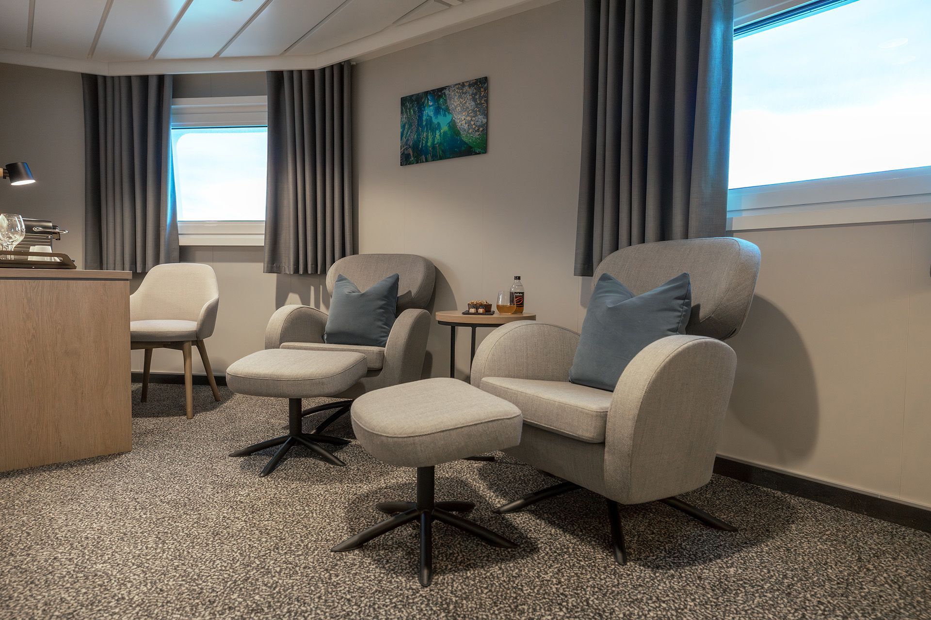 Loungebereich in der Aft View Mini Suite mit zwei bequemen Sesseln und Fußhockern, einem Beistelltisch und großen Fenstern, die natürliches Licht und Meerblick bieten.