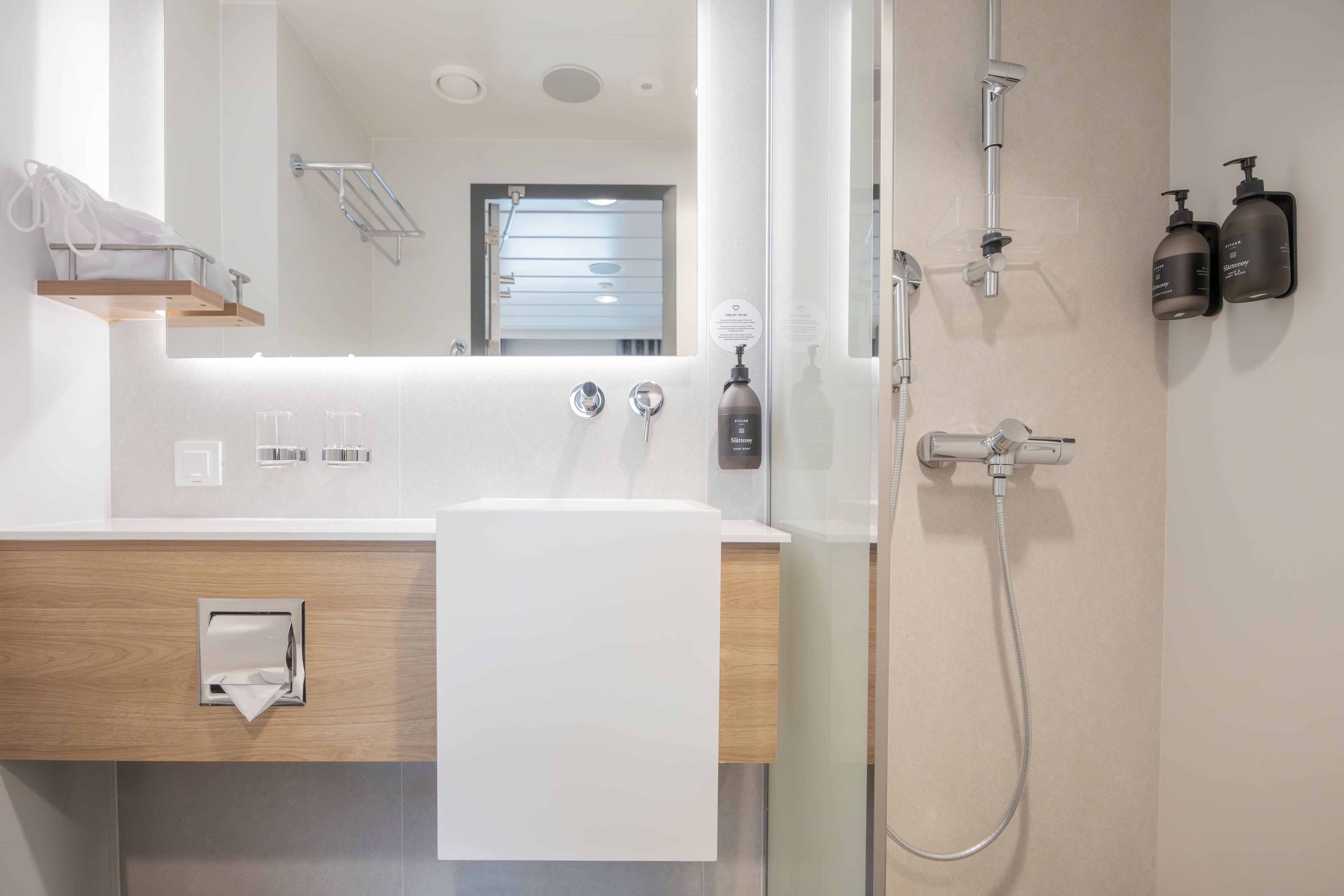 Modernes Badezimmer in der Aft View Mini Suite mit begehbarer Dusche, freihängendem Waschbecken mit beleuchtetem Spiegel, hochwertigen Pflegeprodukten, Ablagefächern und hellen, modernen Oberflächen.