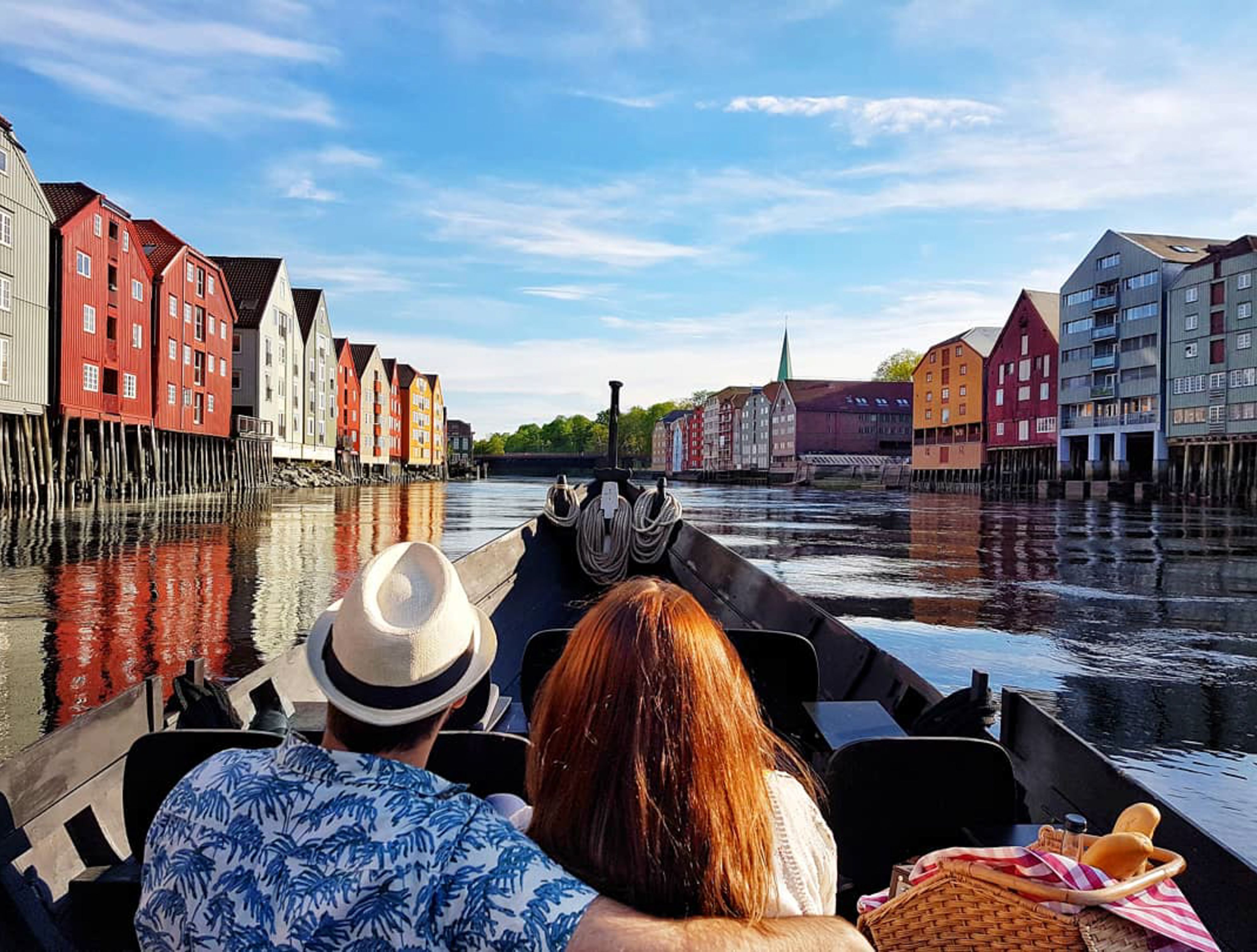 Paar mit Picknickkorb auf einer Bootstour mit Blick auf die farbenfrohen Nidelva-Speicherhäuser in Trondheim, Norwegen