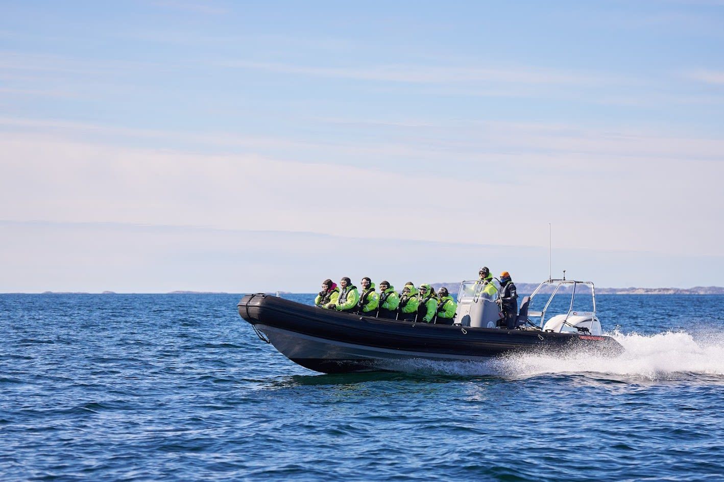 Guests wearing insulated flotation suits speed across the sea on a RIB boat along Bodø’s Arctic coastline, with clear blue skies, open water, and distant islands on the horizon.