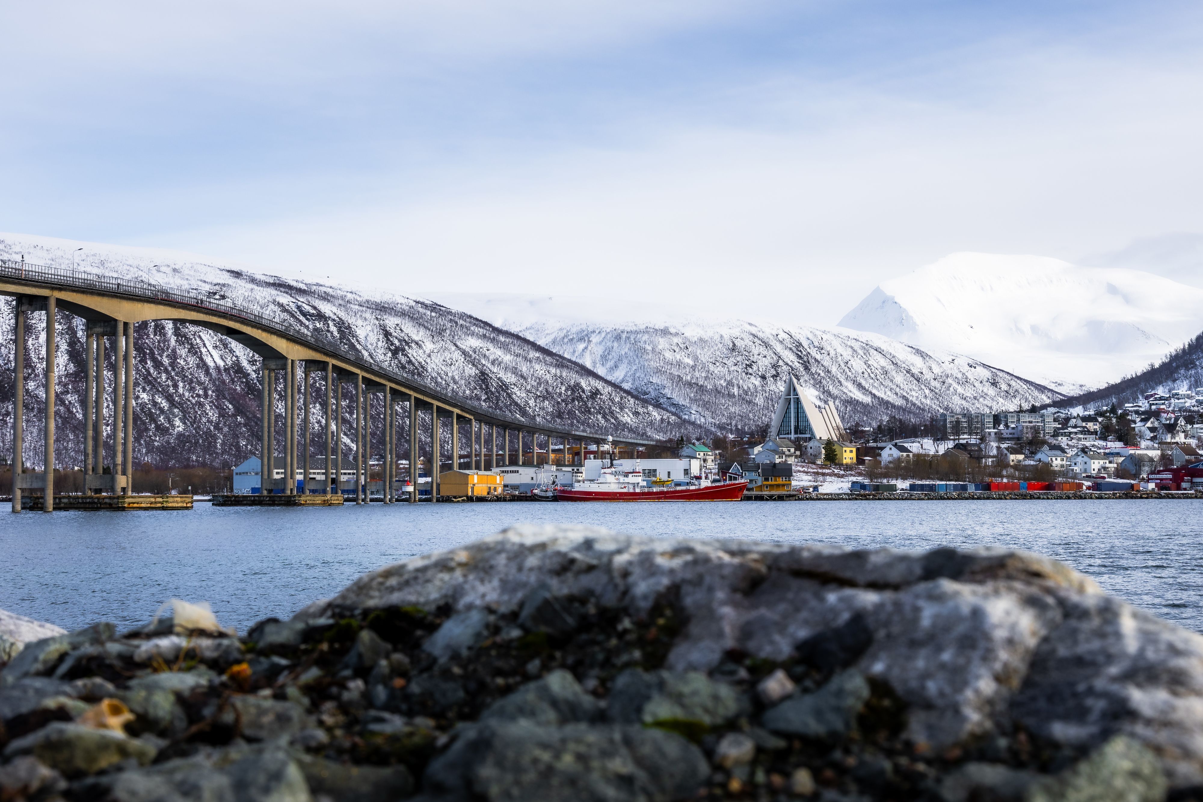 View of the Tromsø Bridge and the iconic Arctic Cathedral with snow-covered mountains and the harbour in the background.