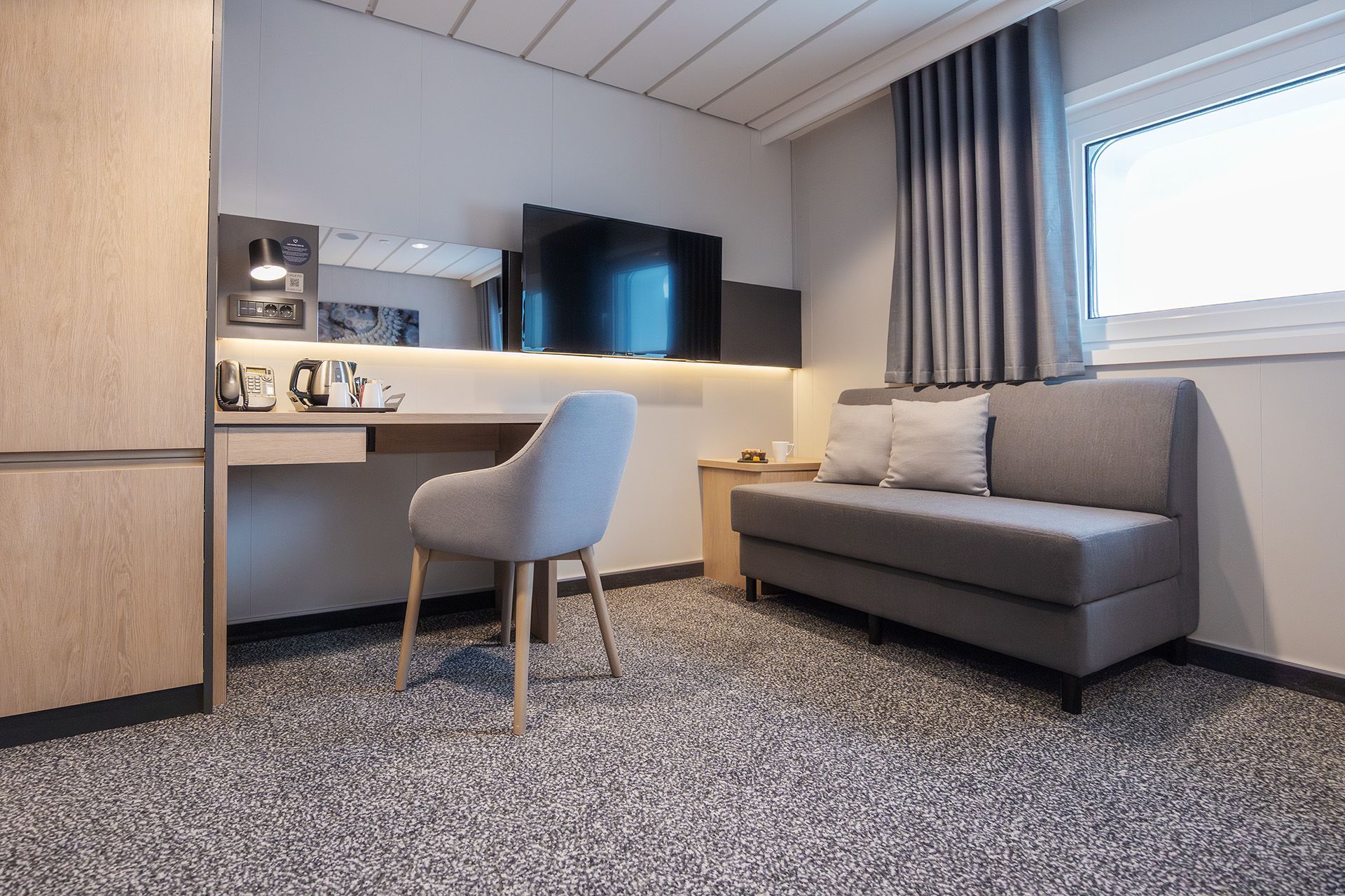 Aft View Superior Double cruise cabin with seating area, sofa, desk, TV, coffee station, and panoramic aft-facing window.