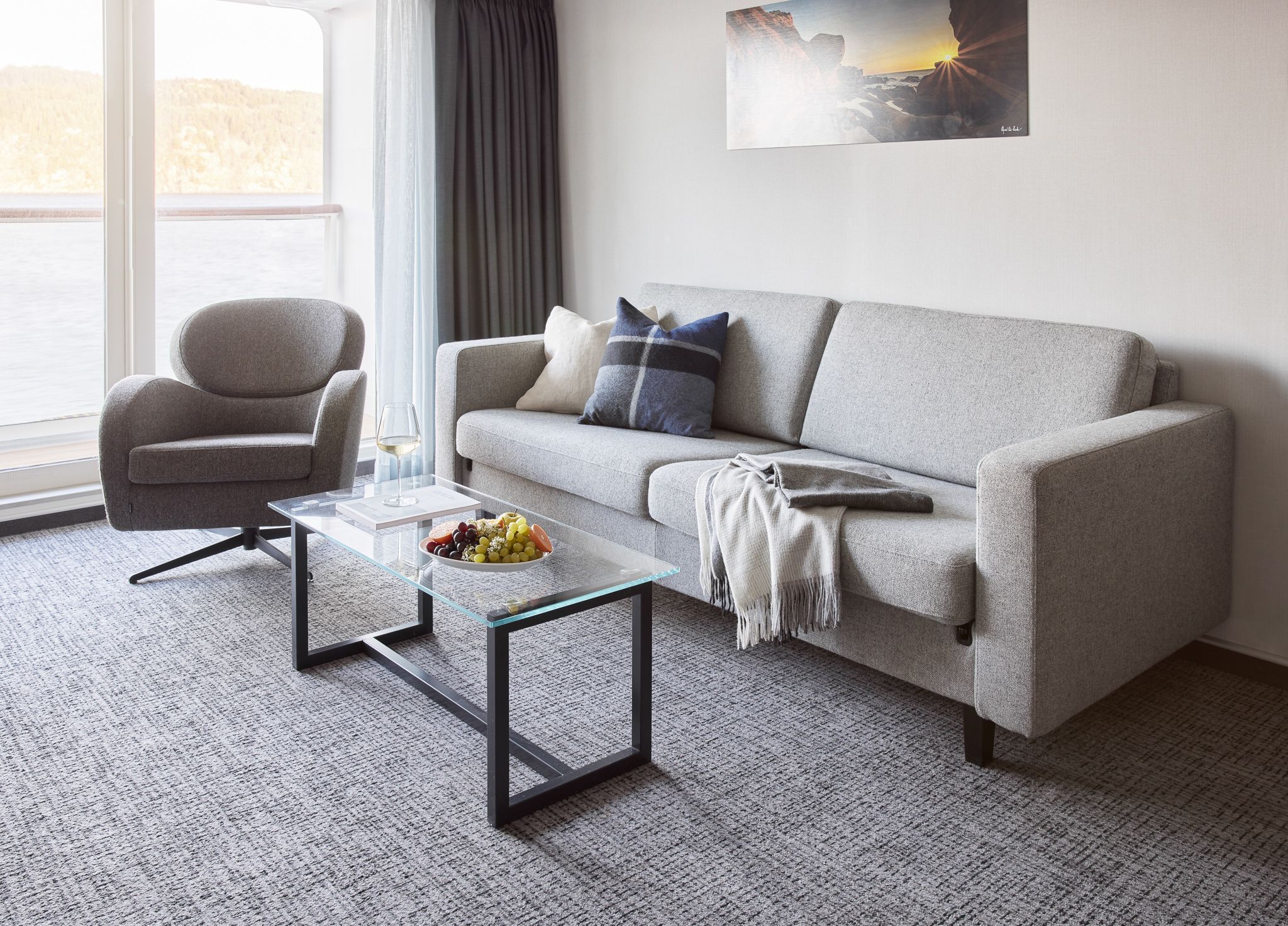 Modern suite living area with a sofa, armchair, glass coffee table, floor-to-ceiling windows opening to a private balcony, and sea views.