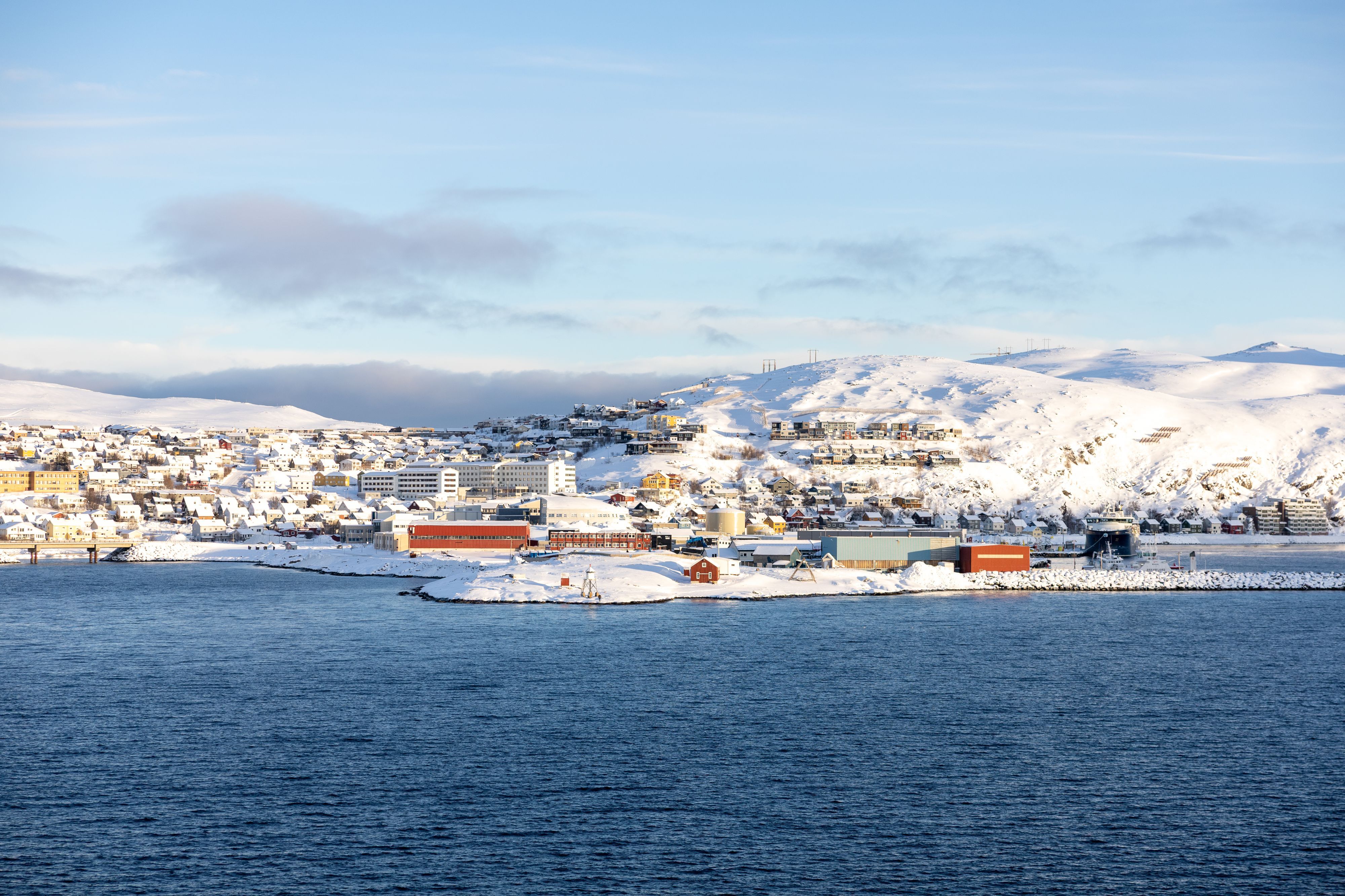 Snødekt kystby Hammerfest sett fra sjøen, med hus i åssiden under en blå vinterhimmel, Norge