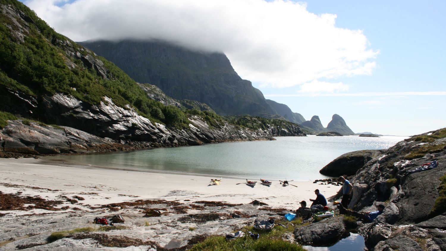 Versteckter weißer Sandstrand auf den Lofoten in Norwegen, mit Seekajaks am Ufer, kristallklarem arktischem Wasser und einer dramatischen Bergkulisse, die eine ruhige Küstenbucht umgibt