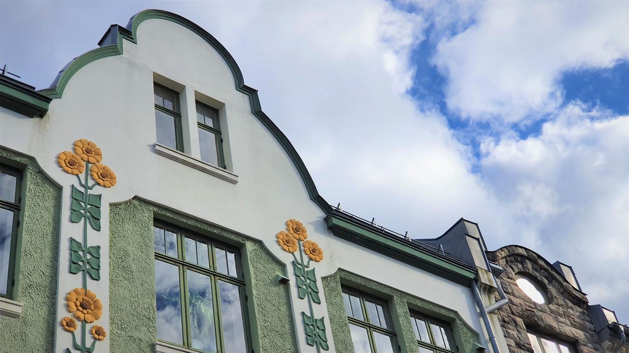 Nahaufnahme eines historischen Jugendstilgebäudes in Ålesund mit geschwungenen Giebeln, dekorativen floralen Motiven und pastellgrünen Verzierungen unter einem hellen, wolkenverhangenen Himmel.