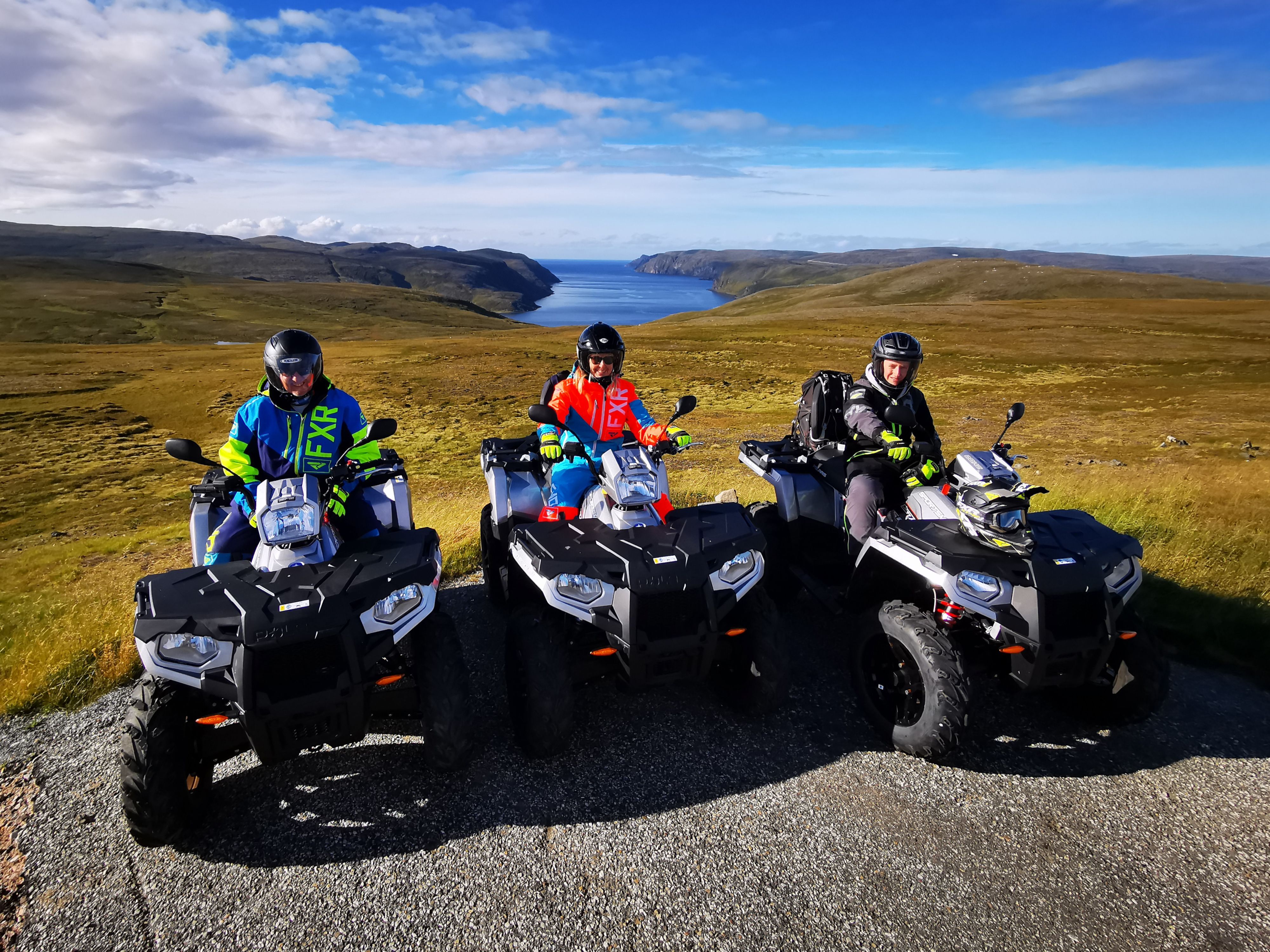 Drei Fahrer auf Quads halten an einem malerischen Aussichtspunkt in der Arktis, von dem aus sie über die sanft geschwungene Tundra und einen tiefblauen Fjord blicken, mit weit offenen Landschaften, die sich auf dem Weg zum Nordkap bis zum Horizont erstrecken.