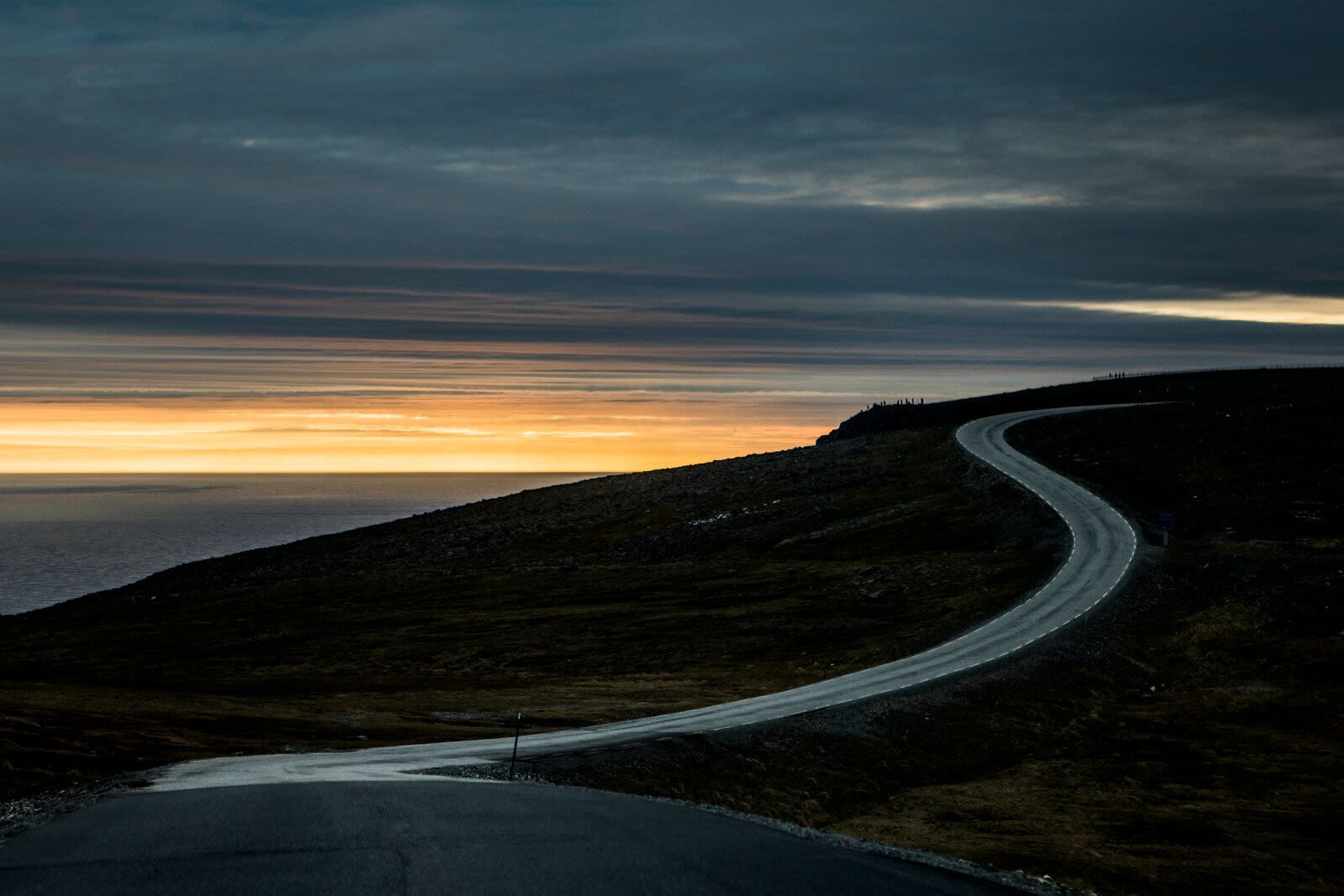 En svingete vei krysser det karrige Nordkapp-platået mot Polhavet, der et gyllent skjær bryter frem under dramatiske skyer i horisonten.