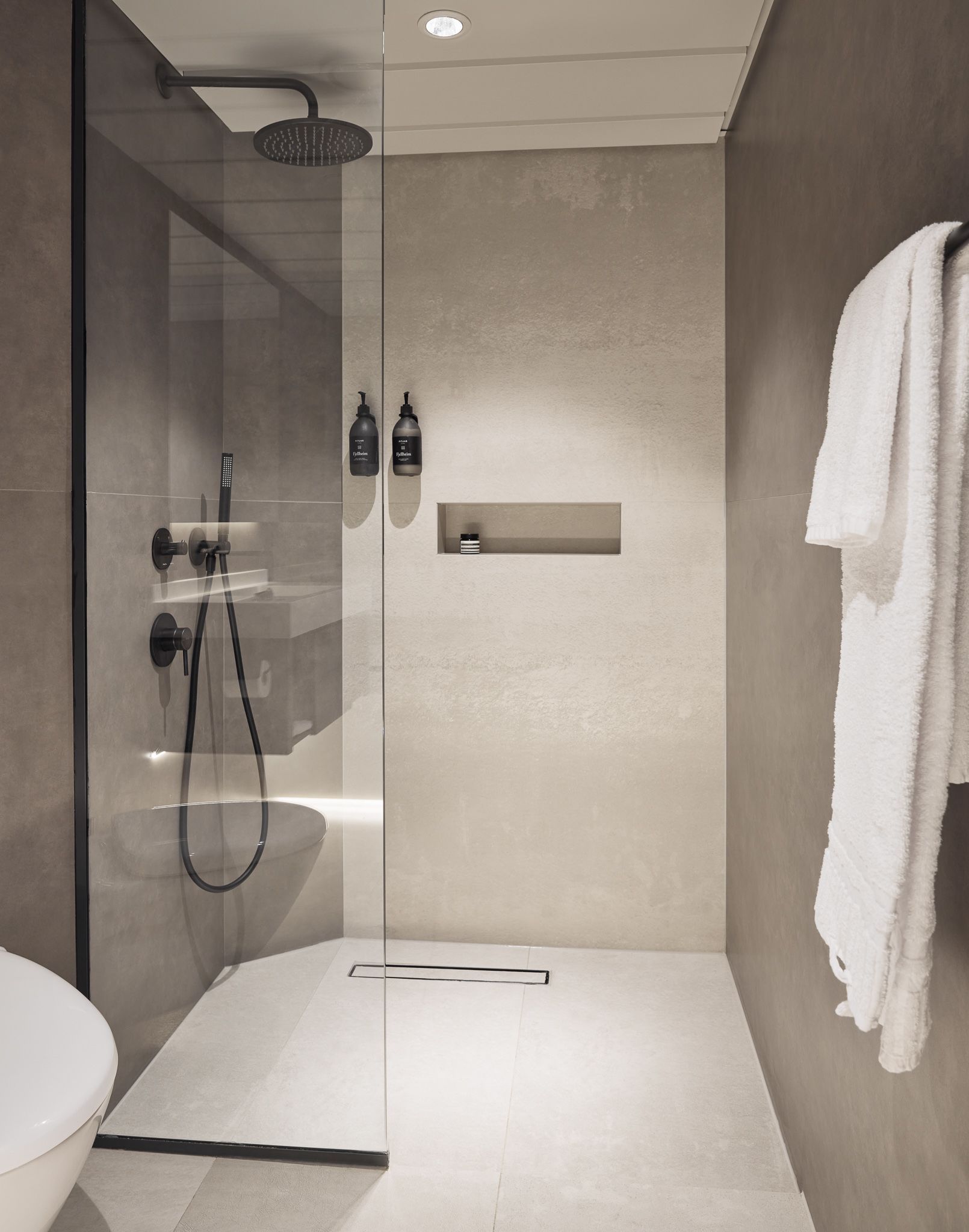 Begehbare Regendusche mit Glasabtrennung, mattschwarzen Armaturen, integrierter Ablage und wandmontierten Pflegeprodukten in einem großzügigen, modernen Badezimmer.