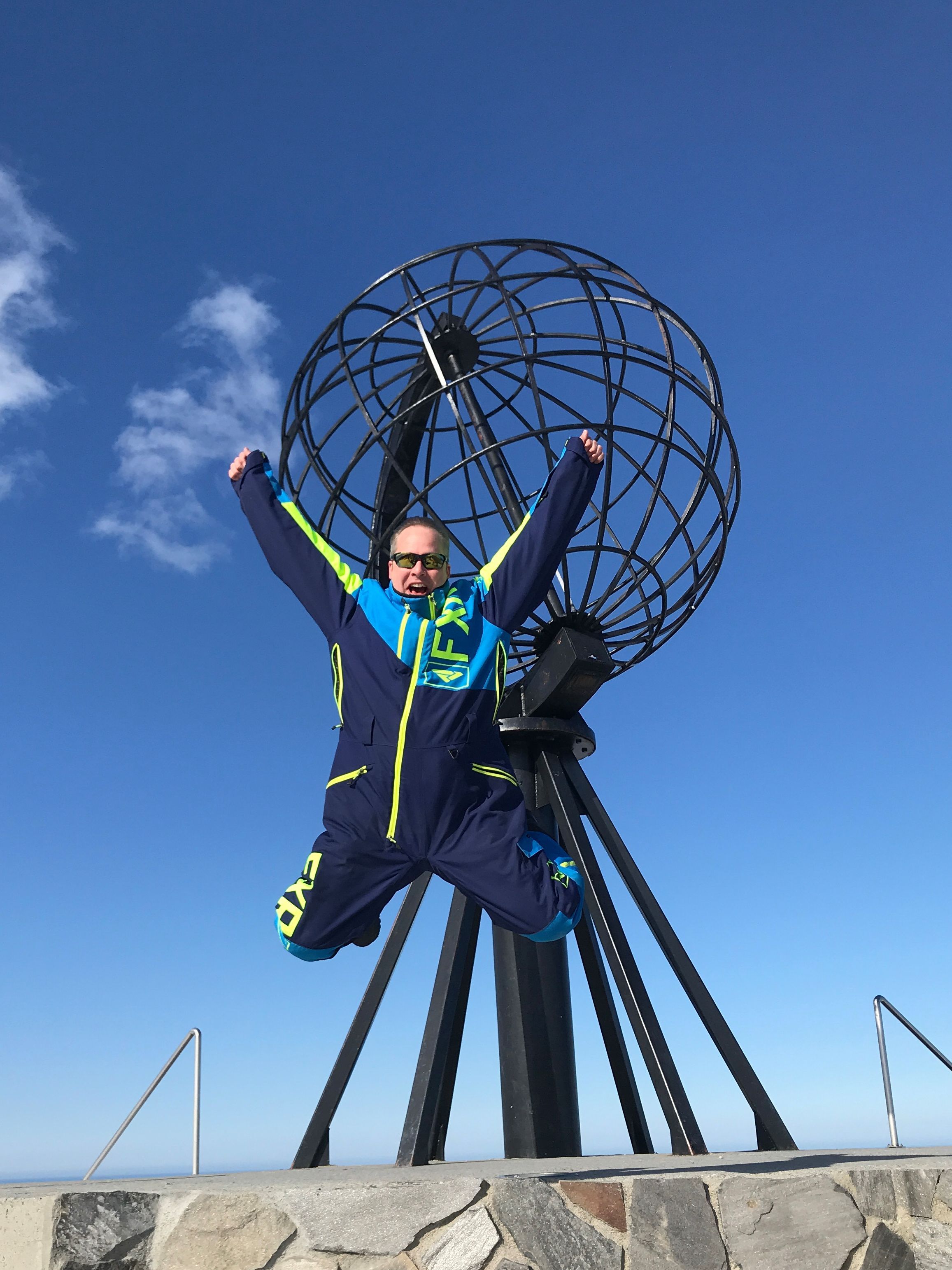 Ein Besucher springt mit erhobenen Armen vor dem ikonischen Nordkap-Globusdenkmal in den klaren blauen arktischen Himmel und feiert an einem der nördlichsten Wahrzeichen Europas.